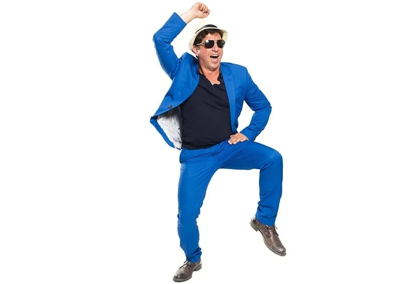 Ein lachender Mann in blauer Anzug, Sonnenbrille, Hut und Tanzposen vor weißem Hintergrund.