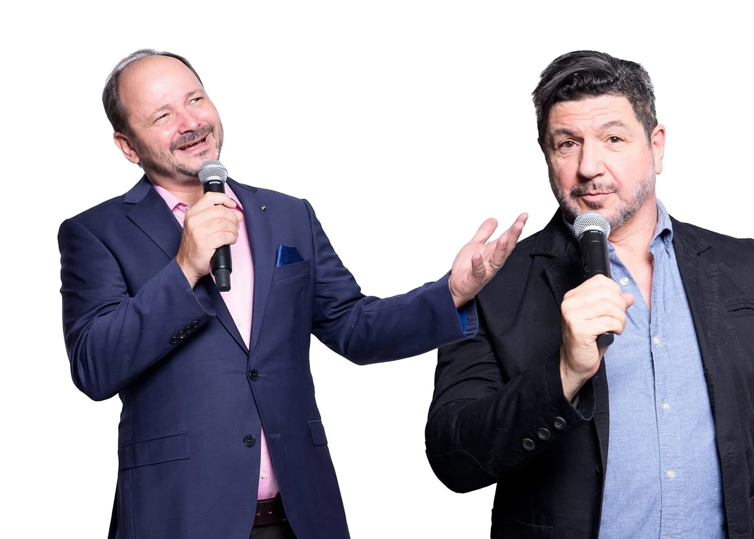 Zwei Männer sprechen auf einer Bühne, einer von ihnen ist lächelnd und trägt einen dunklen Anzug mit rosa Hemd, der andere schaut ernst und trägt eine dunkle Jacke mit blauer Hemd, beide halten Mikrofone.