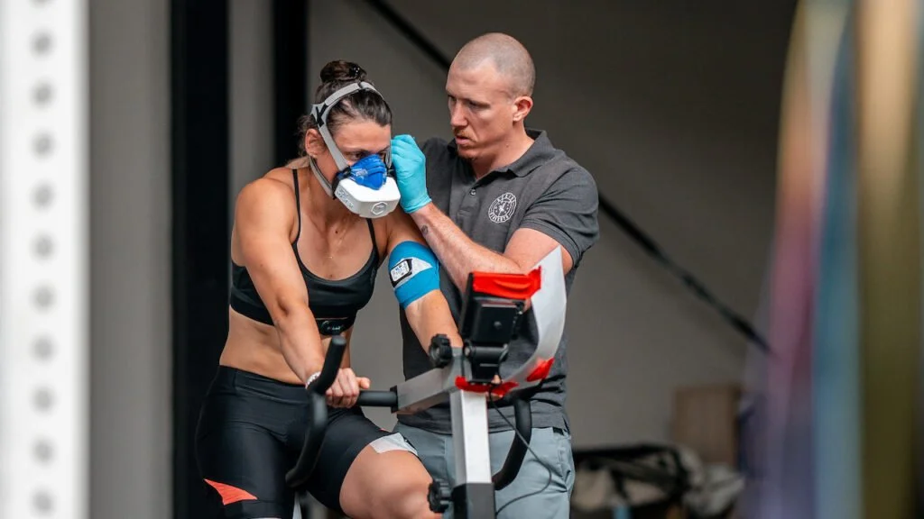 A woman wearing a breathing mask and sports attire is fitted with an EMS suit on her upper body while a man adjusts the straps, with a sports chair in the background.