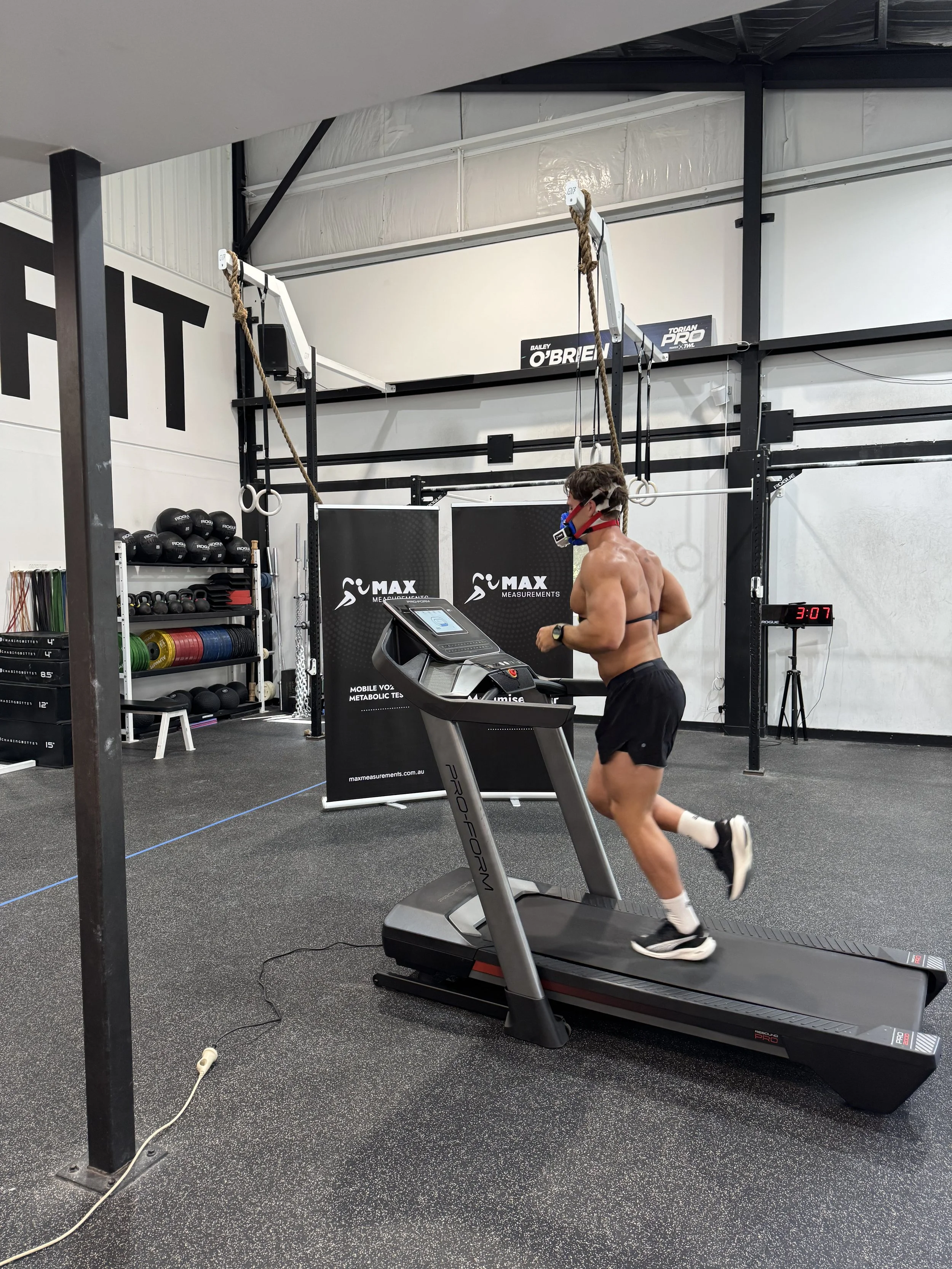 A shirtless man running on a treadmill in a gym, wearing a face mask, glasses, and running shoes.