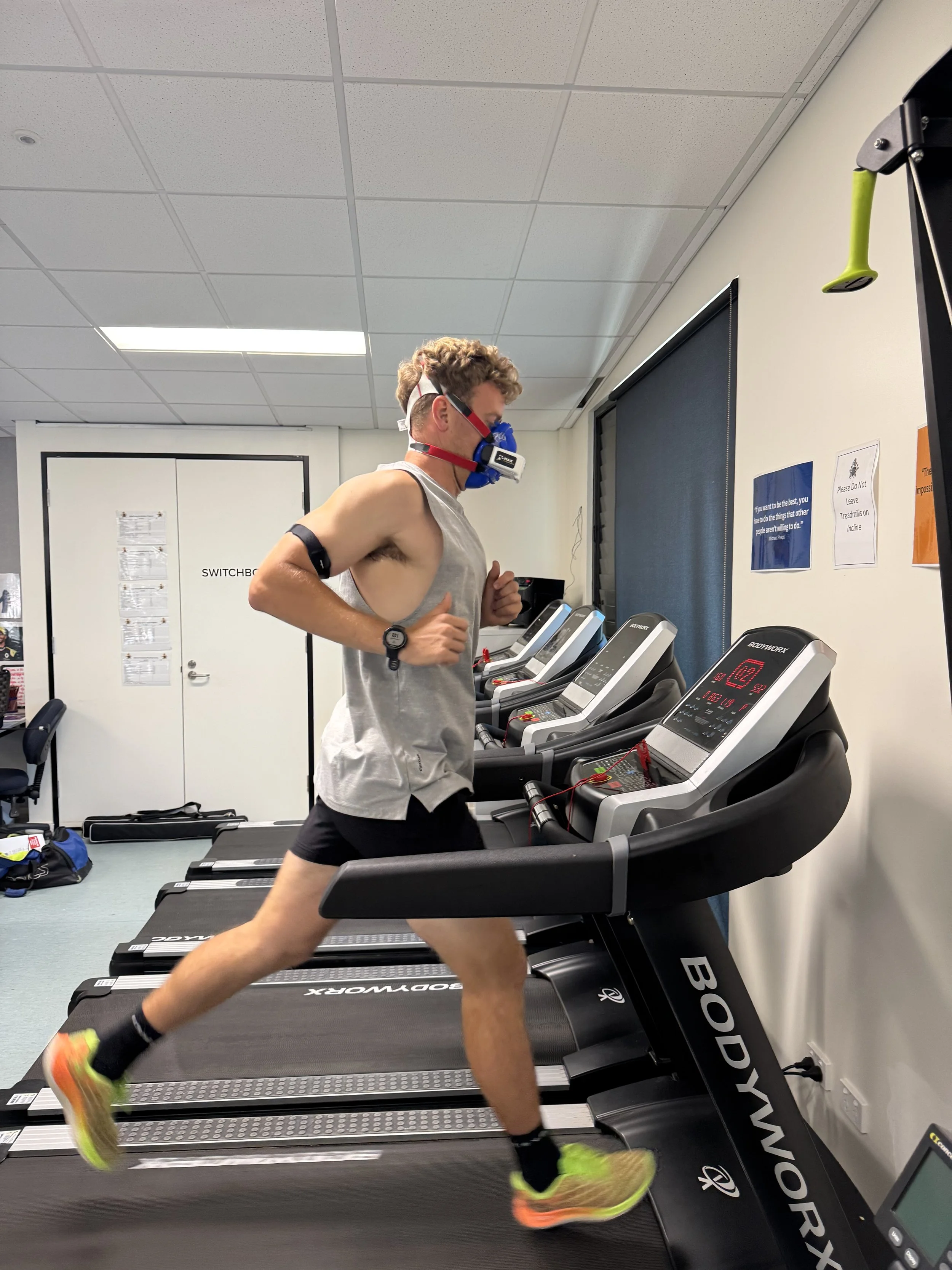 A man running on a treadmill in a gym, wearing a gray sleeveless shirt, black shorts, bright shoes, a fitness tracker, and a protective face mask with a headgear.