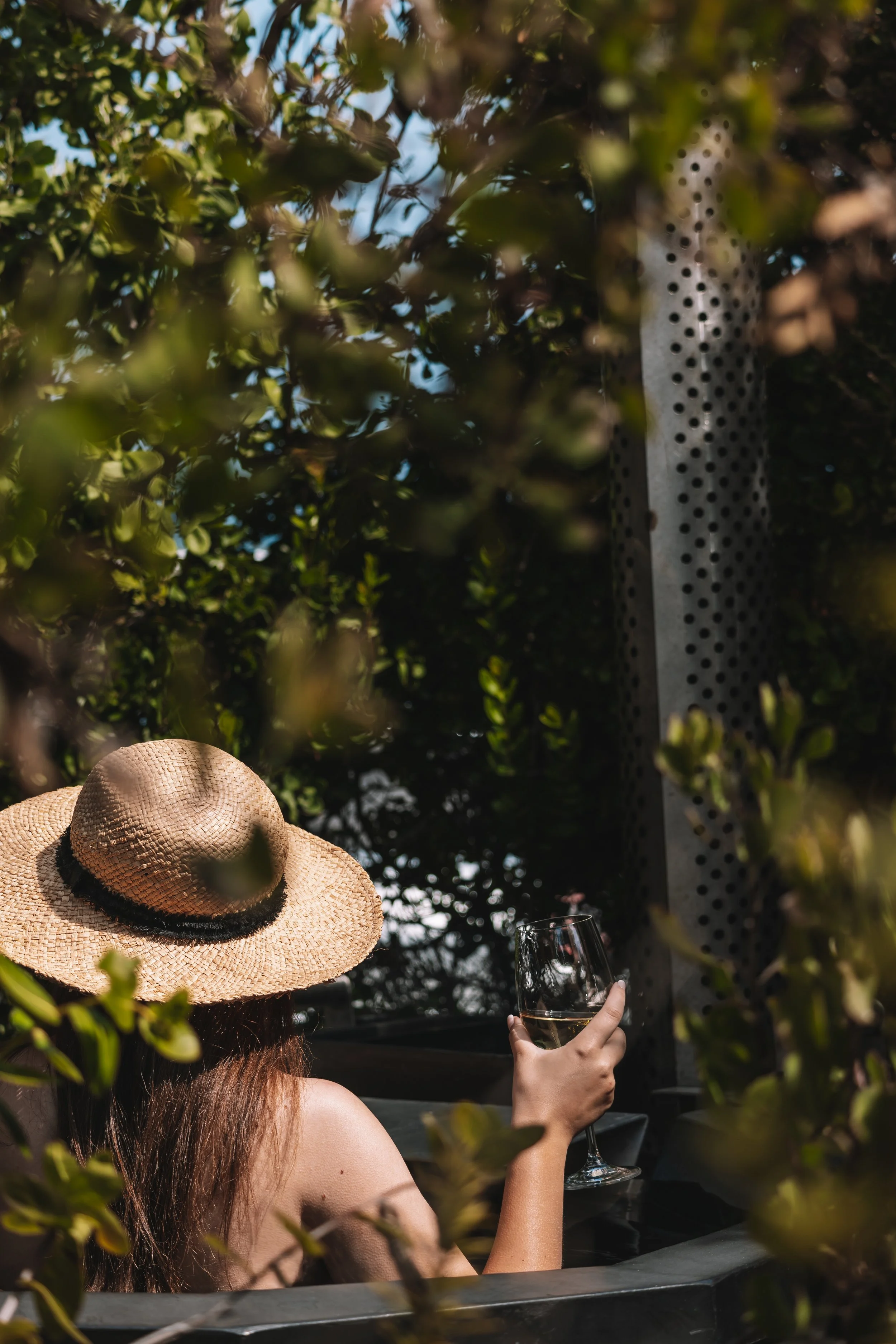 A woman with long hair wearing a wide-brimmed straw hat is sitting outdoors, holding a glass of white wine, surrounded by green foliage.