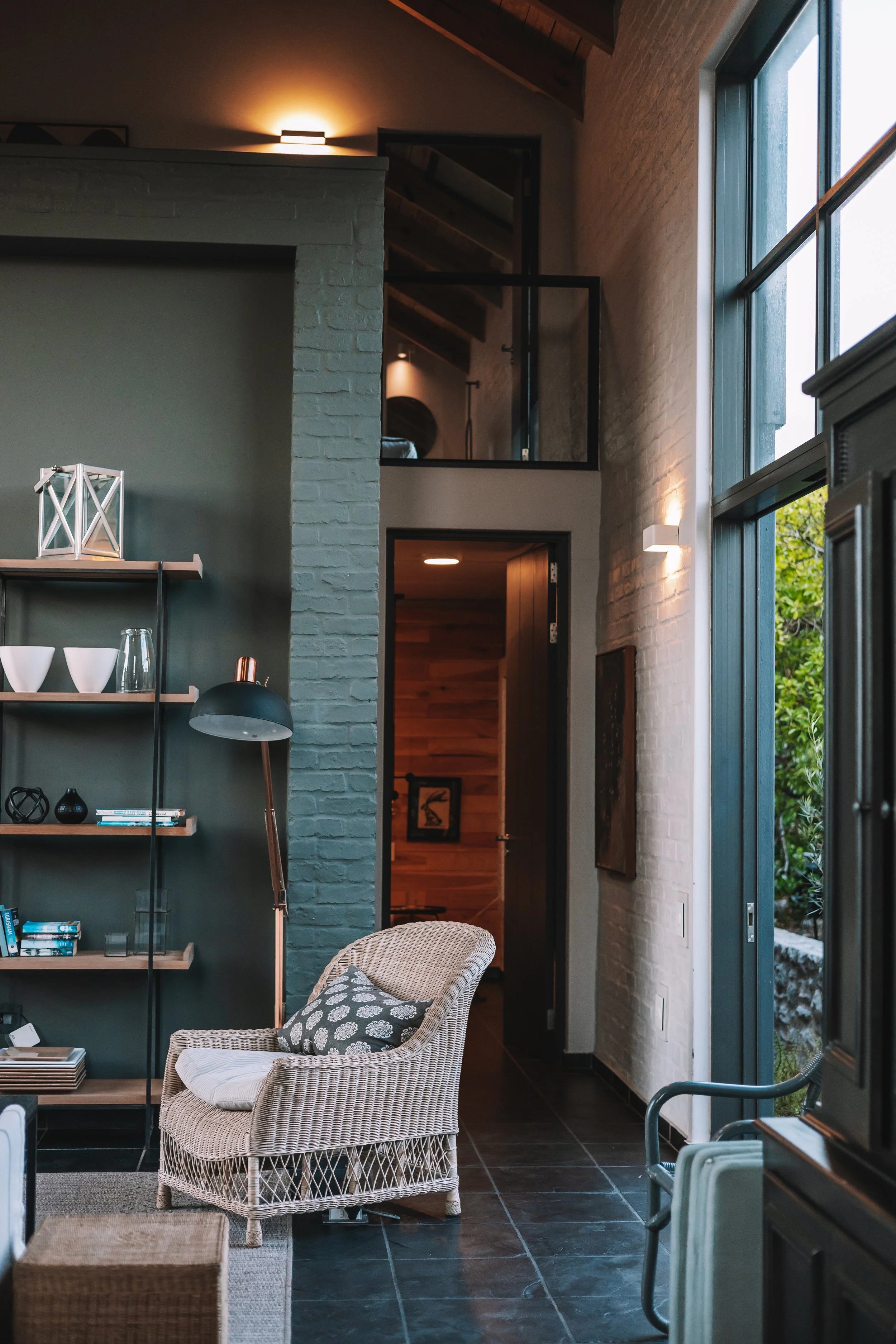 Interior of a modern living space with a wicker armchair, bookshelf, and large windows showing greenery outside.
