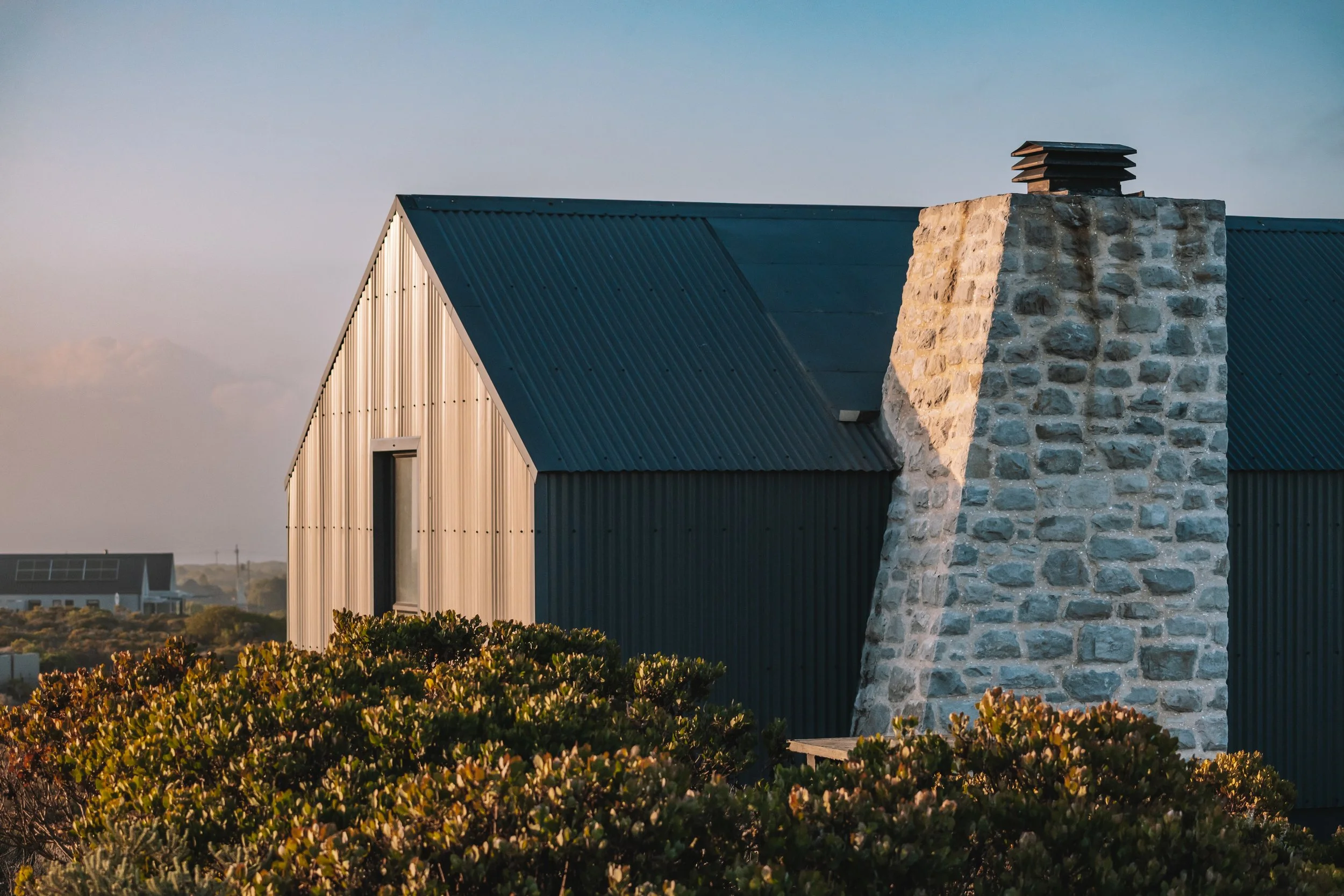 A modern house with a blue metal roof and a stone chimney surrounded by greenery during sunset.