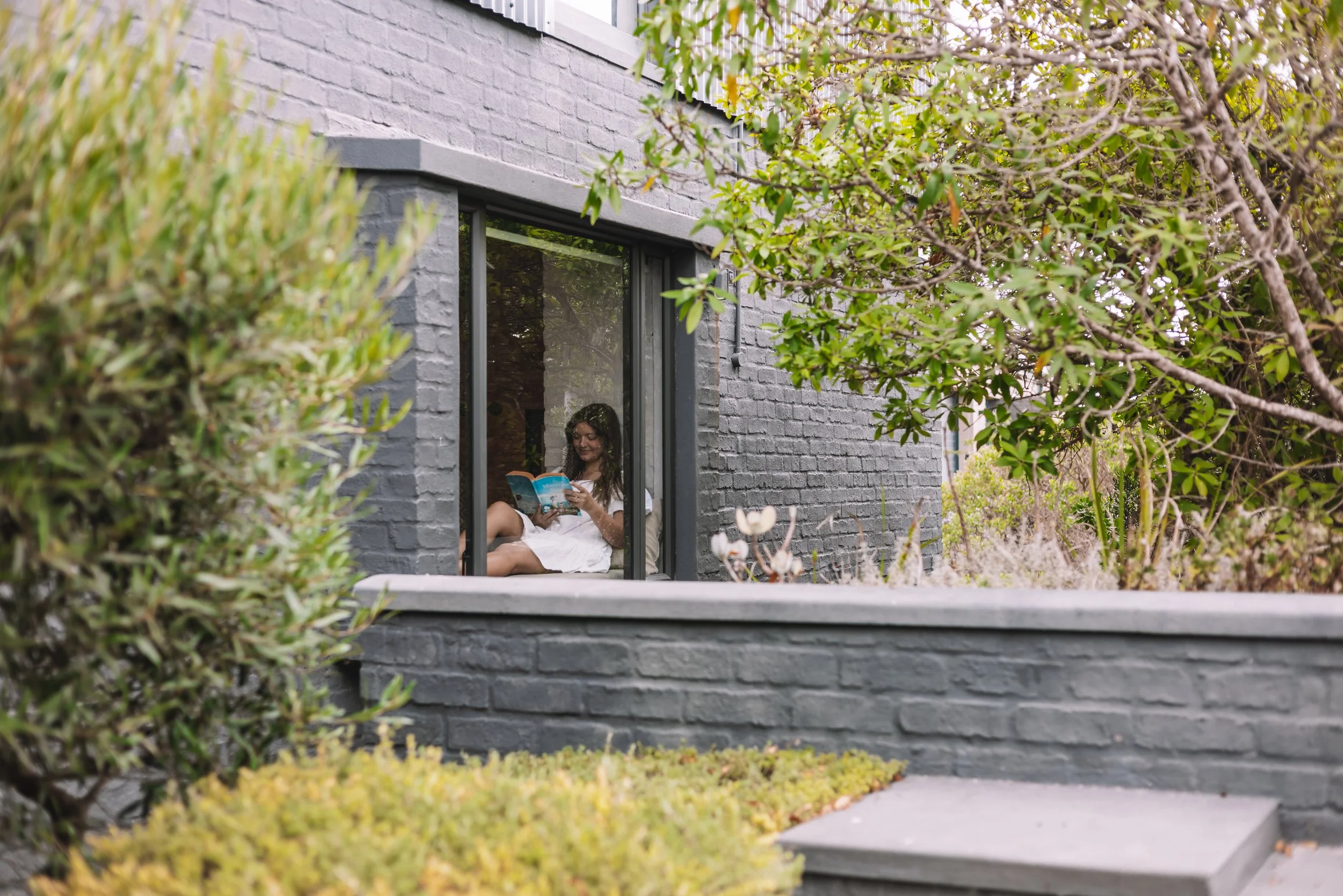 A woman in a white dress sitting on a window ledge, reading a book, with greenery and bushes around a gray brick building.