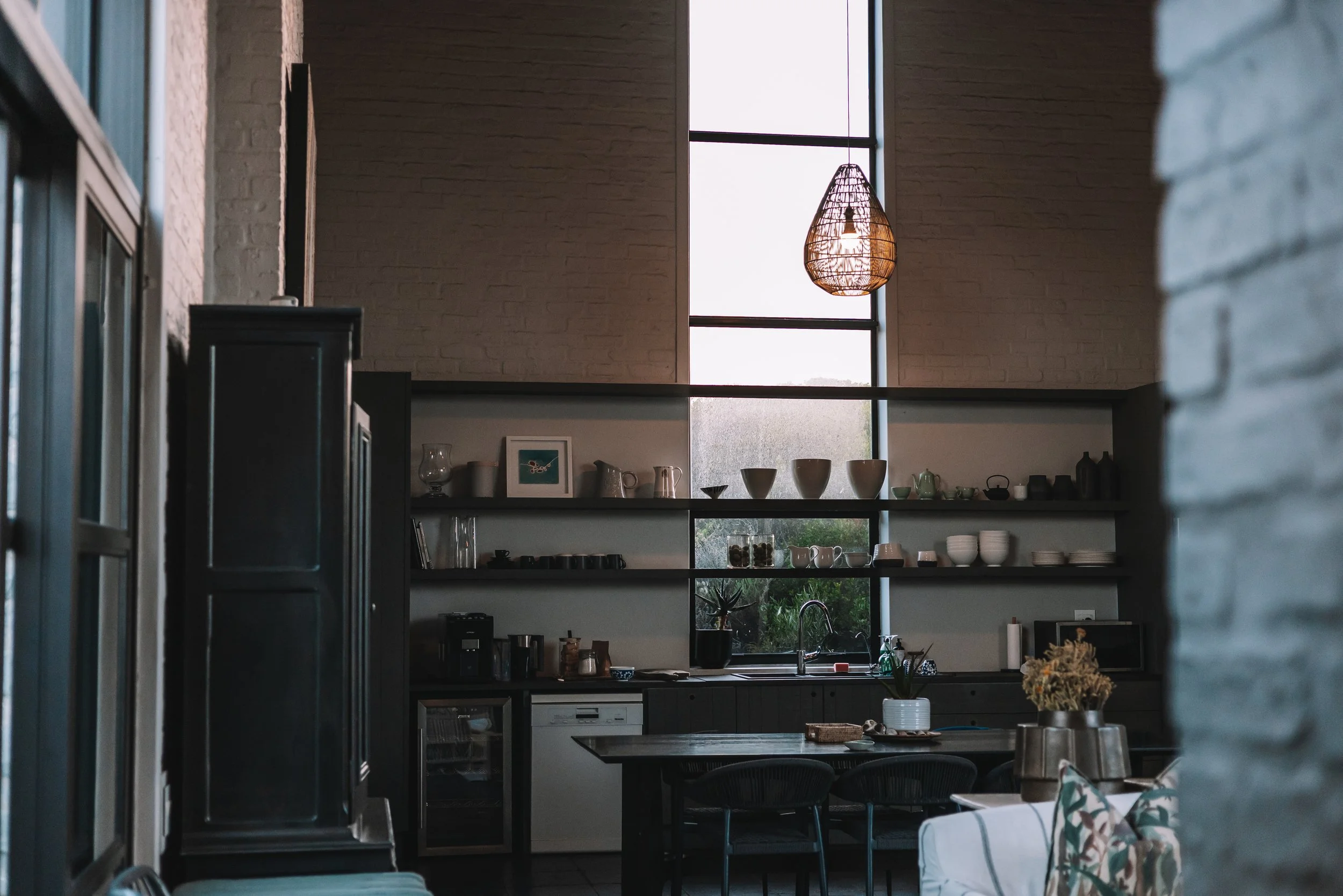 Interior view of a modern kitchen with open shelving, large window, hanging wicker light fixture, and black cabinetry.