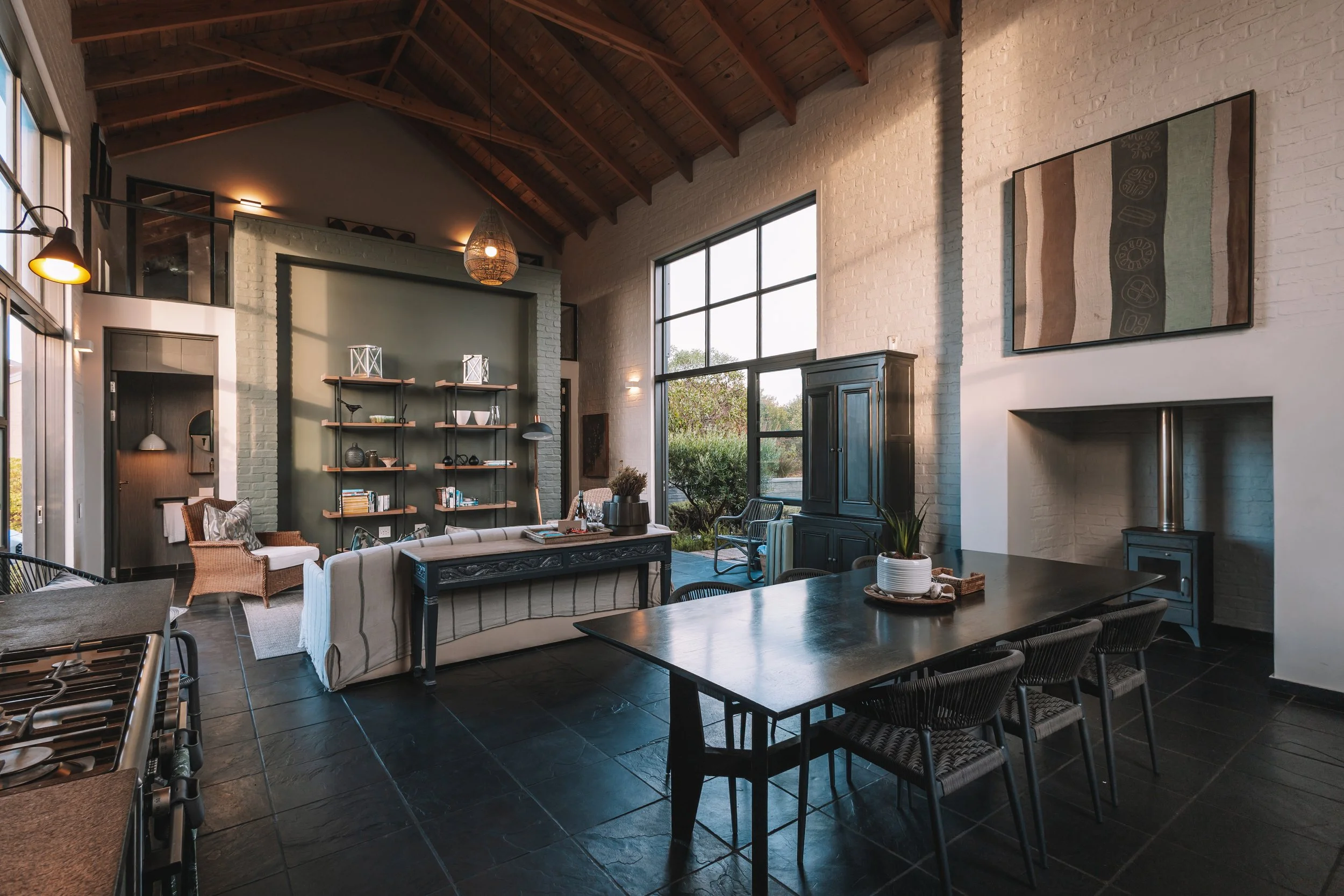 Interior of a modern living room with high wooden ceiling, large windows, a black bookshelf, a dining table with six chairs, a fireplace, and various decorative items.