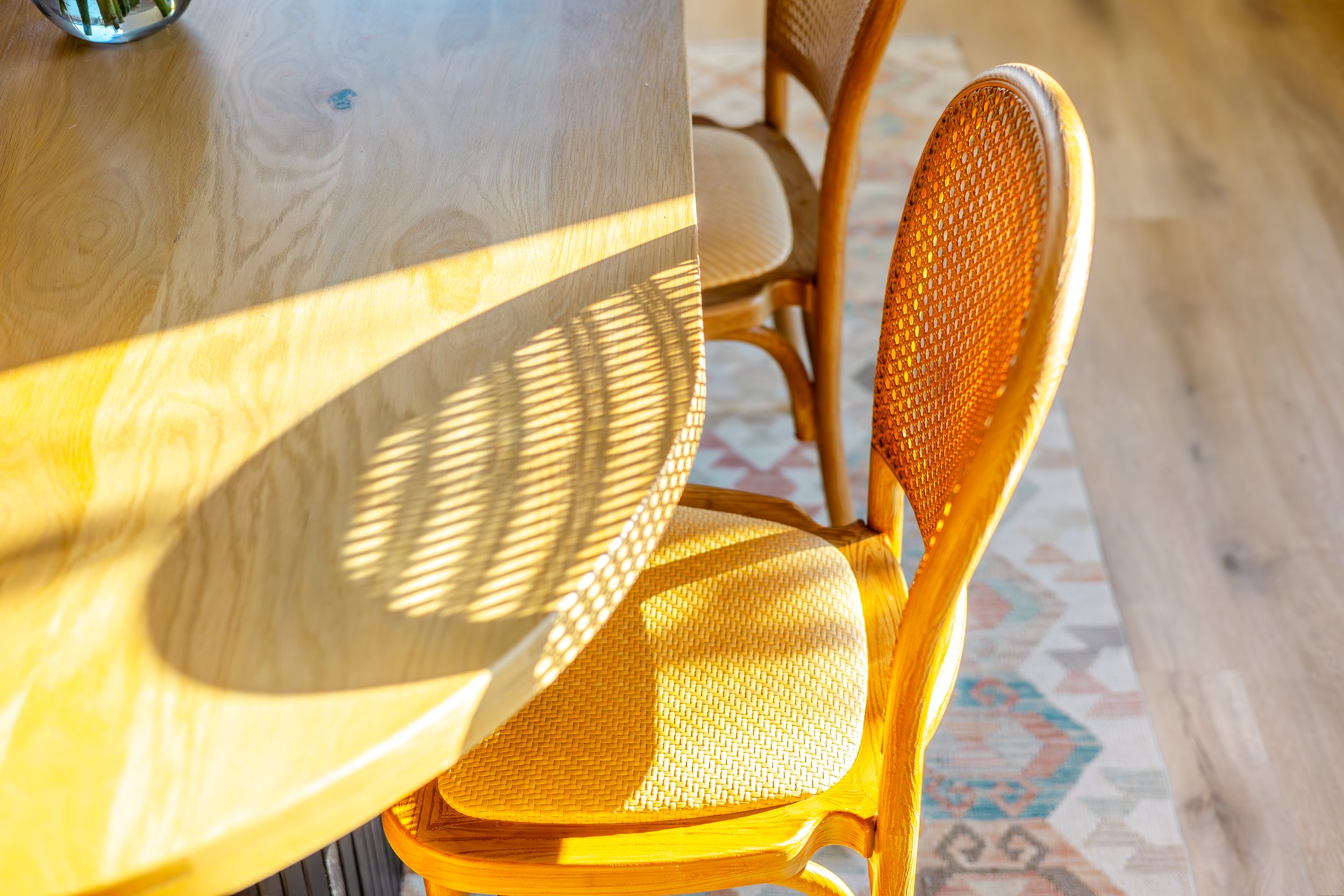 Sunlit wooden dining table with woven cane-backed chairs, partial view of a floor rug with geometric patterns.