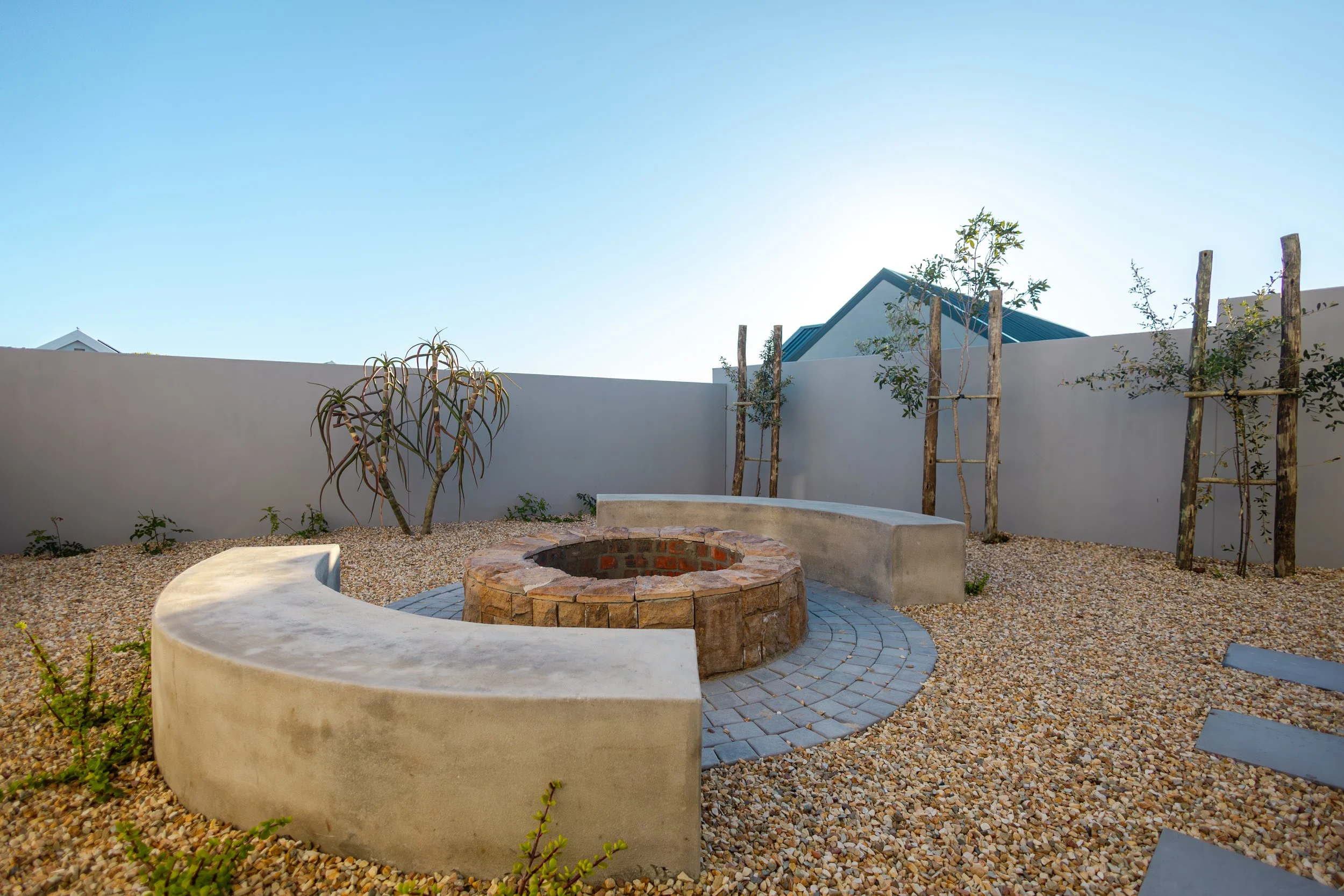 A minimalist backyard with a circular brick fire pit, curved concrete seating, and a gravel ground, surrounded by young trees supported by wooden stakes, with a plain white wall and a clear blue sky in the background.