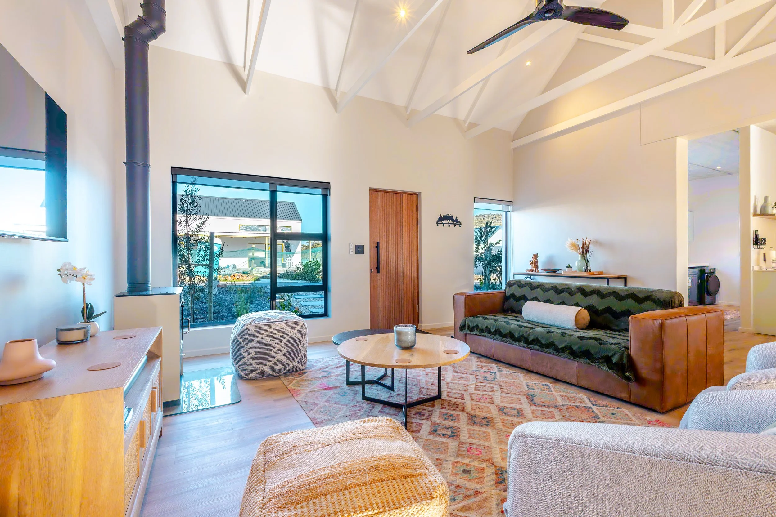 Bright, modern living room with white walls and vaulted ceiling, furnished with a brown leather sofa, a patterned armchair, and a round coffee table on a colorful rug; large windows and a sliding door reveal a garden area outside.