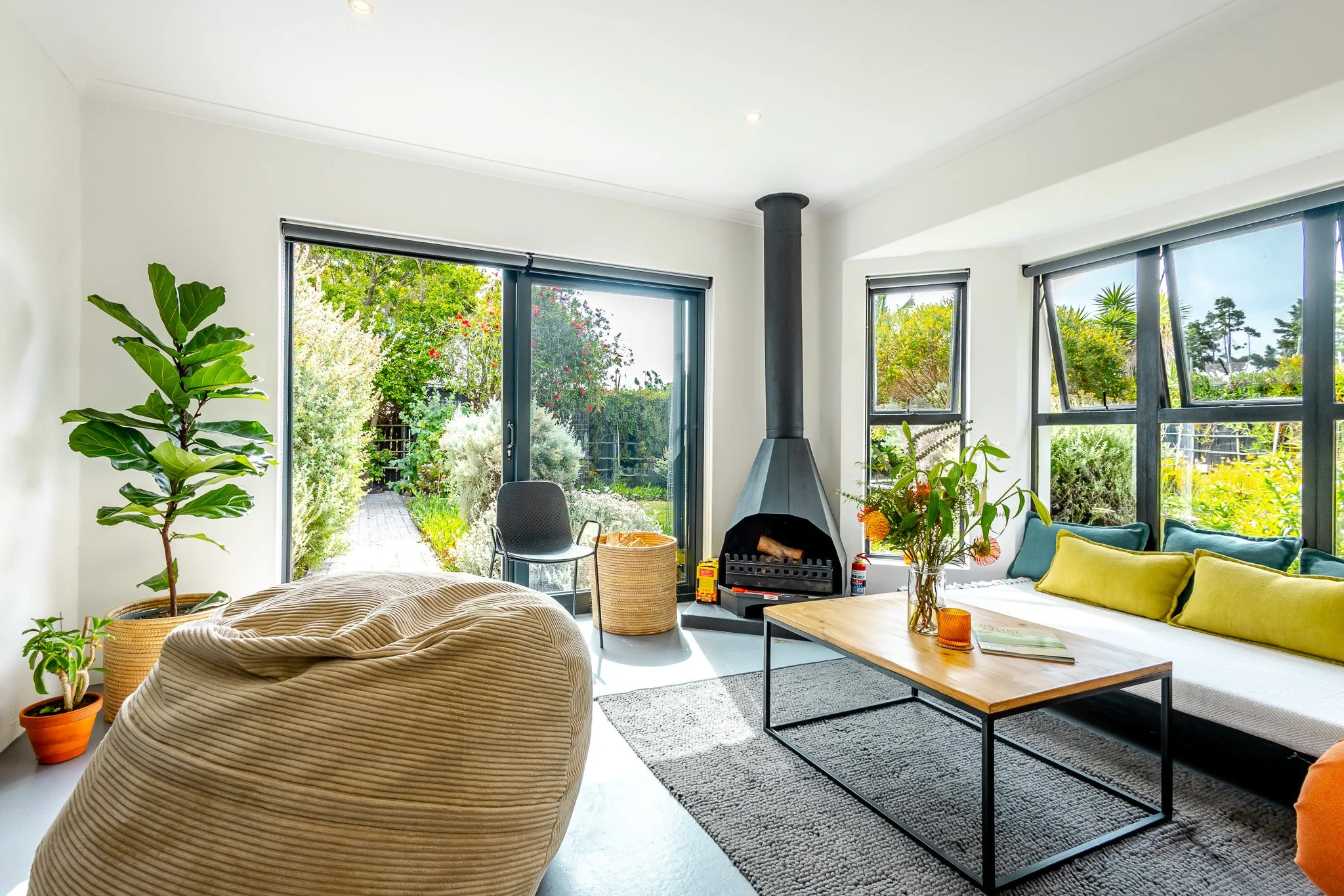 Professional real estate photography of a sun-lit Kommetjie lounge featuring a modern fireplace, beanbag seating, and large glass doors opening to a lush garden.