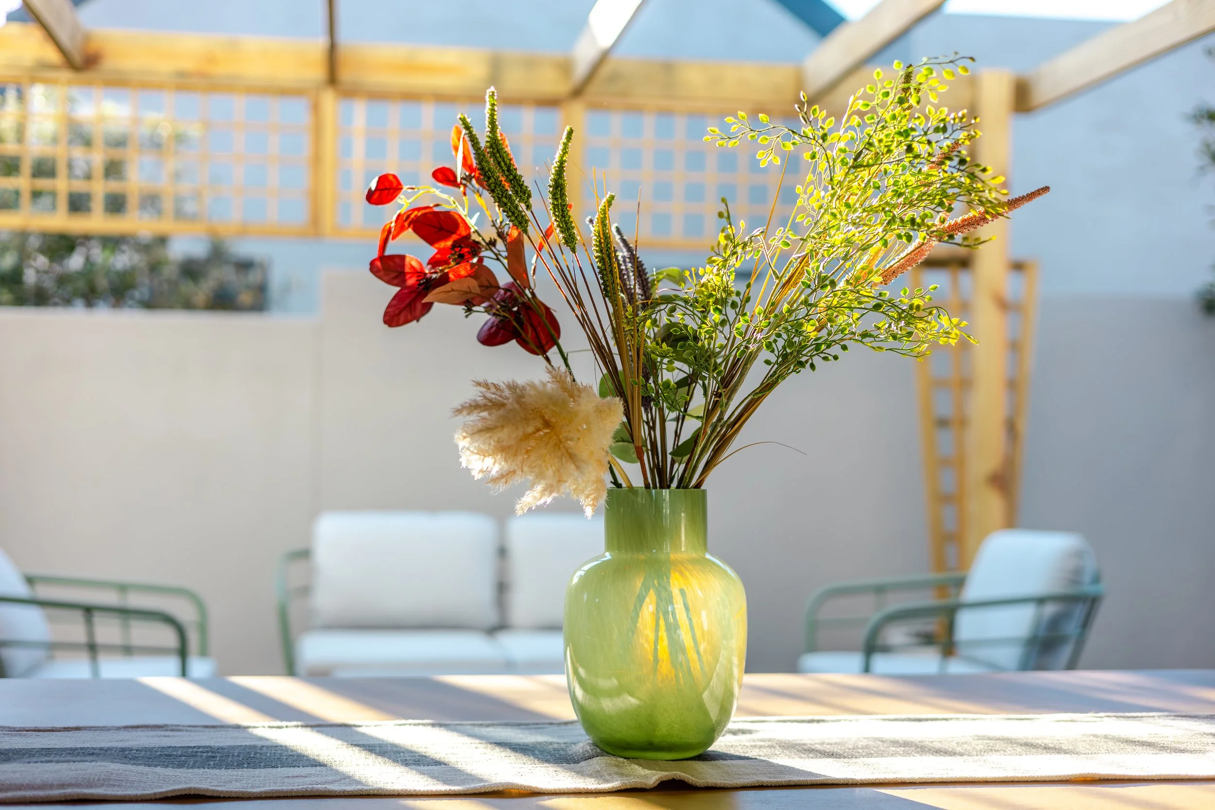 Green vase with assorted flowers and greenery on a table in an outdoor patio.
