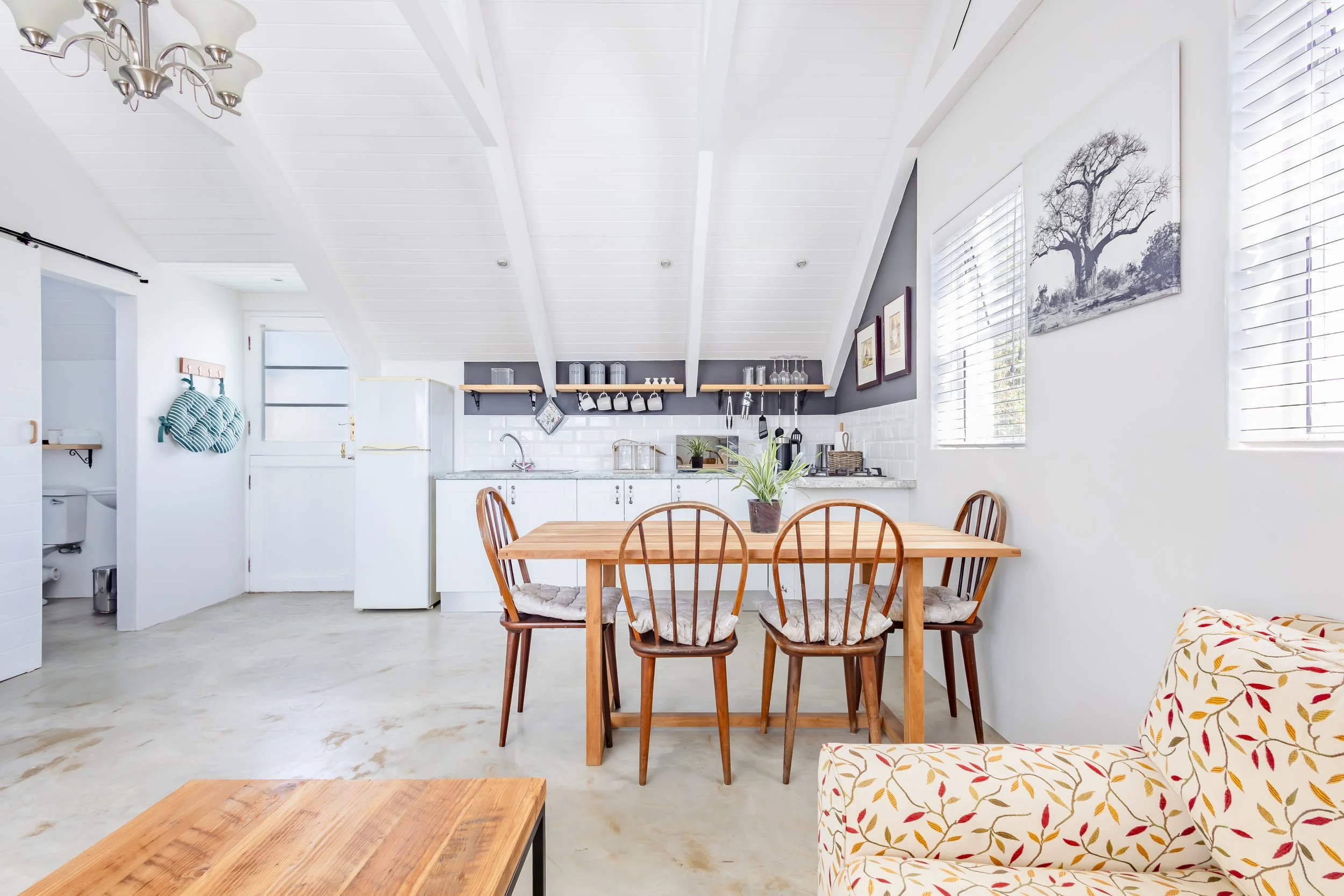 Professional real estate photography of a modern open-plan kitchen and dining area with white vaulted ceilings, wooden furniture, and clean finishes.