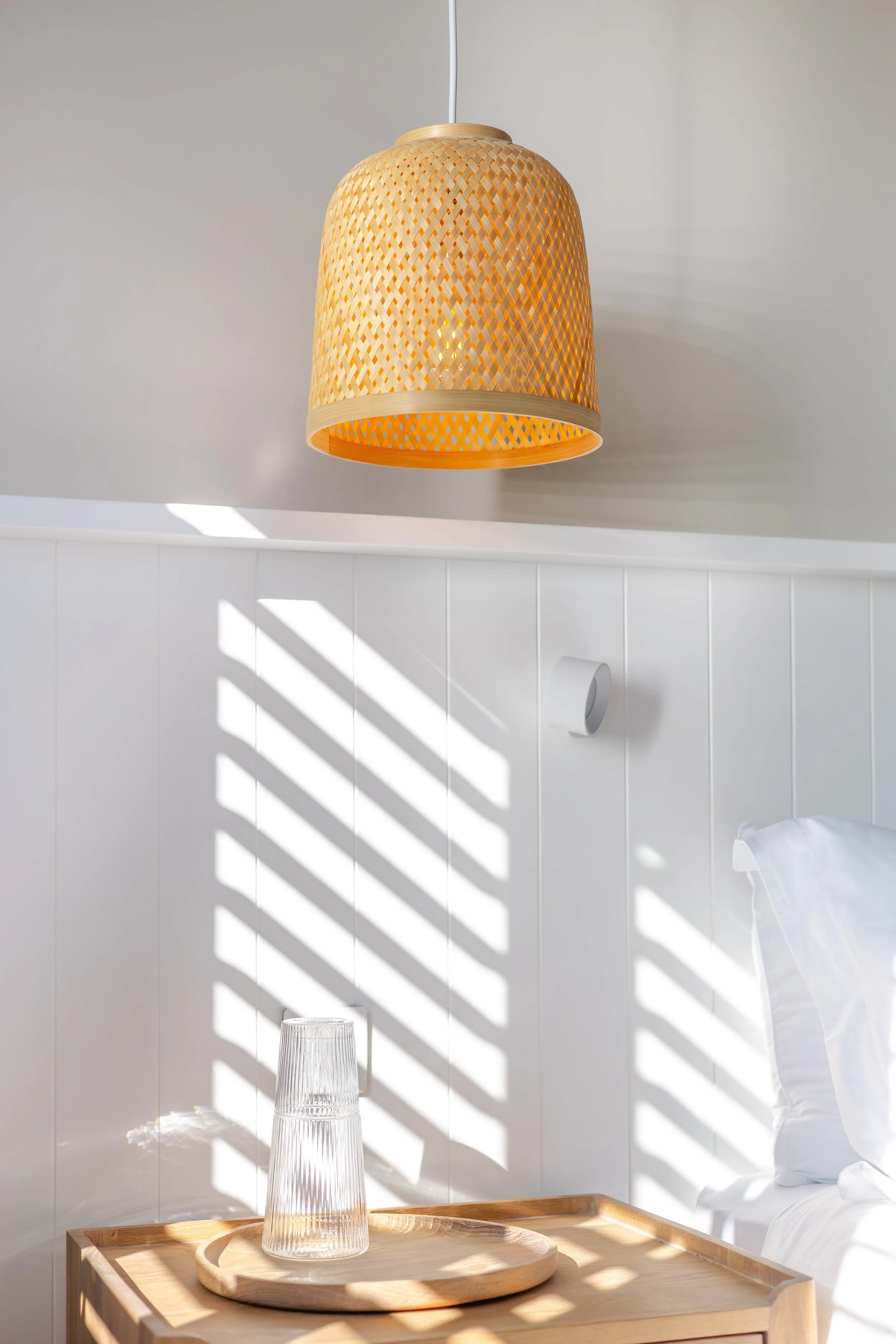 A modern bedroom with a hanging woven pendant lamp, sunlight creating striped shadows on the wall, a wooden tray with stacked glass cups, and white bedding.