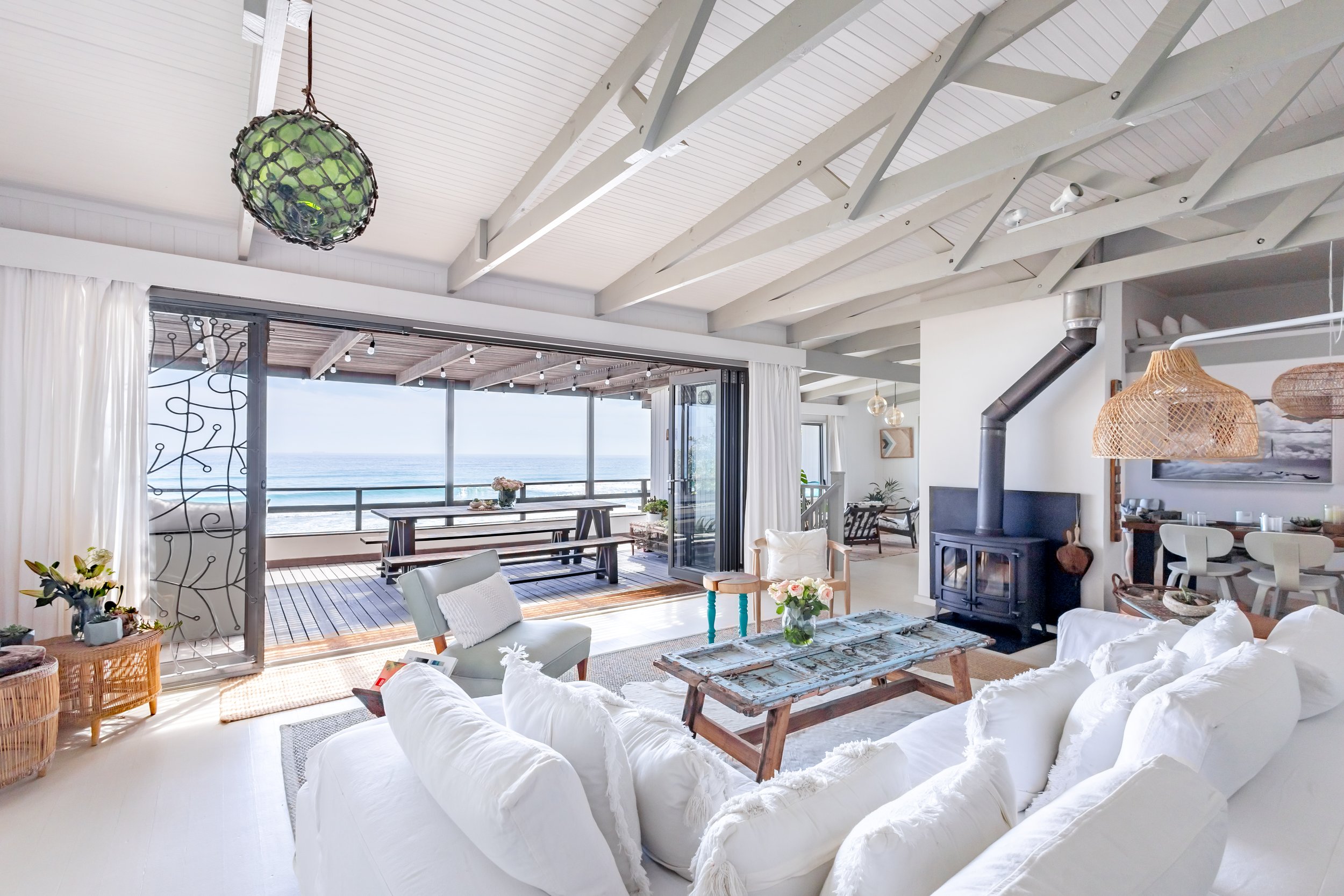 Bright, open-plan living room with white sofas, a rustic fireplace, and floor-to-ceiling glass doors opening to the sea.