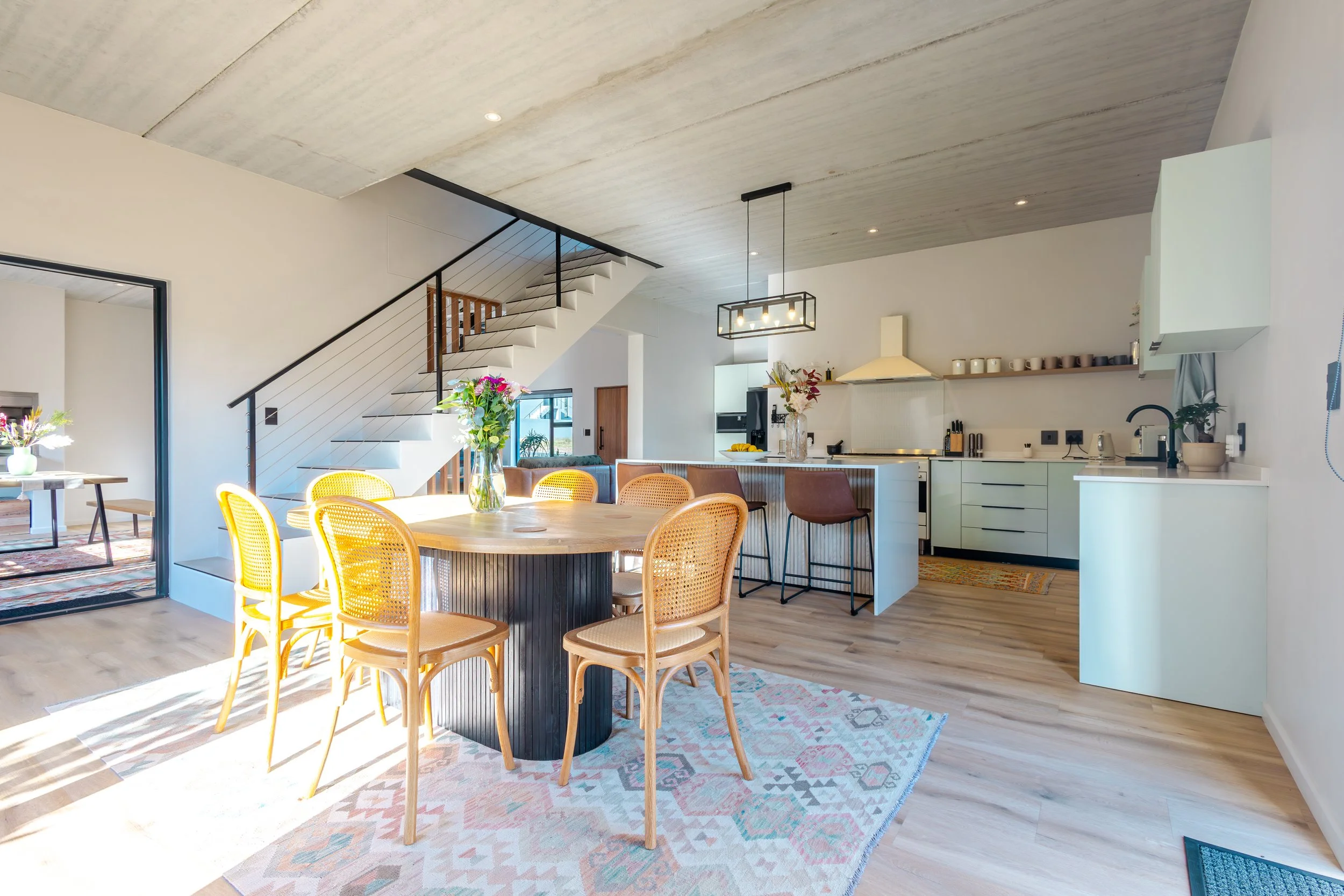 Open-concept modern kitchen and dining area with light wood flooring, white walls, and a concrete ceiling. A round wooden dining table with yellow chairs, a vase with flowers, and a kitchen island with bar stools. Sunlight streams through large windo
