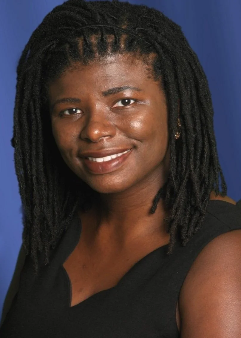 A woman with dark skin tone, wearing a sleeveless black top, smiling with visible teeth, and has shoulder-length black dreadlocks against a blue background.