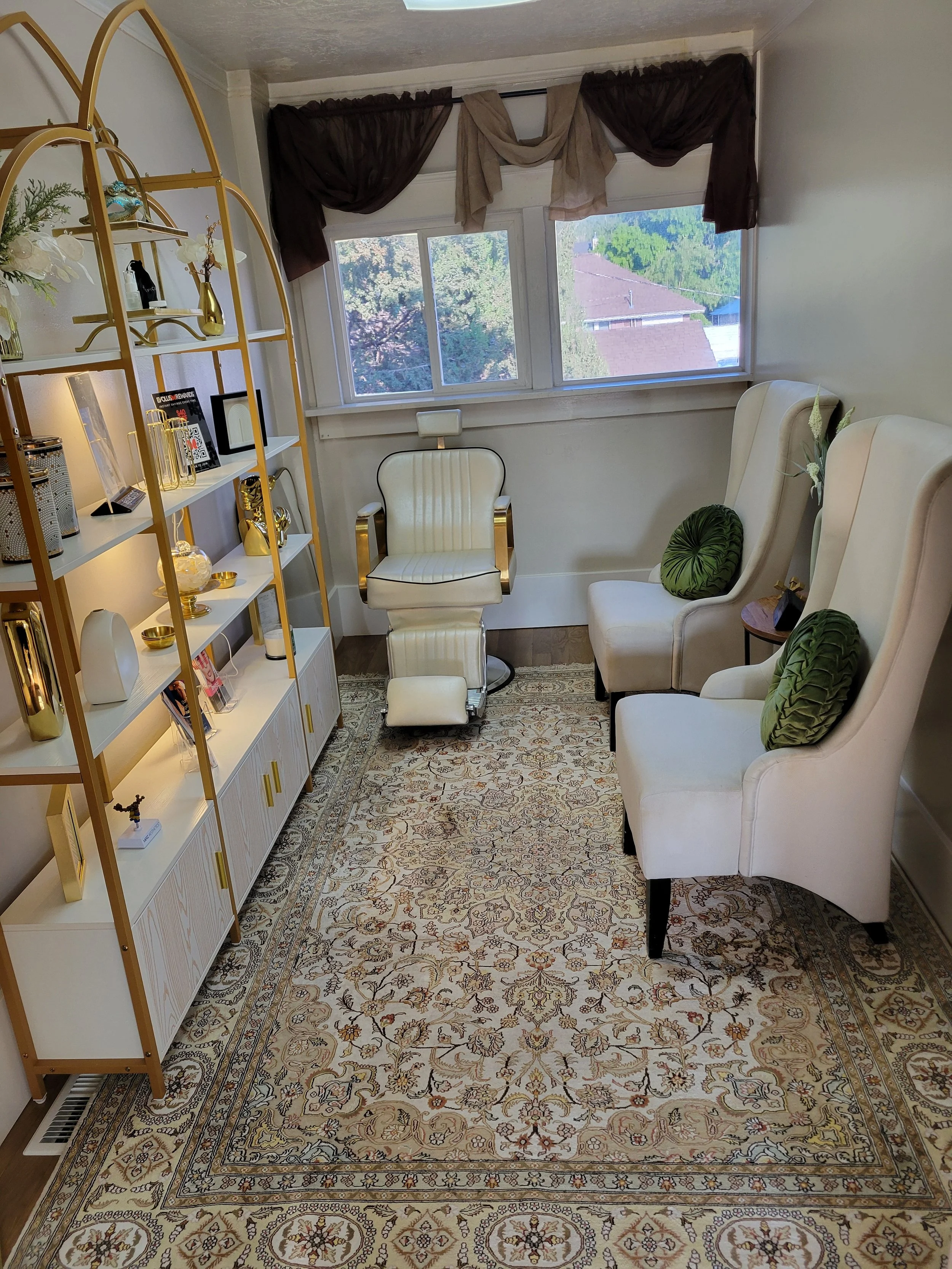 A cozy living room with a large area rug, three white high-backed chairs with green pillows, a gold and white bookshelf filled with decorative items, and a window with dark brown and beige curtains showing a view of trees and rooftops.