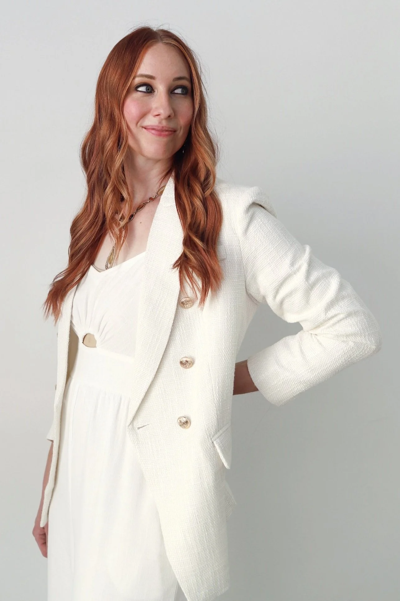 A woman with red hair and light skin wearing a white blazer over a white dress, standing against a light background, looking to the side with a slight smile.