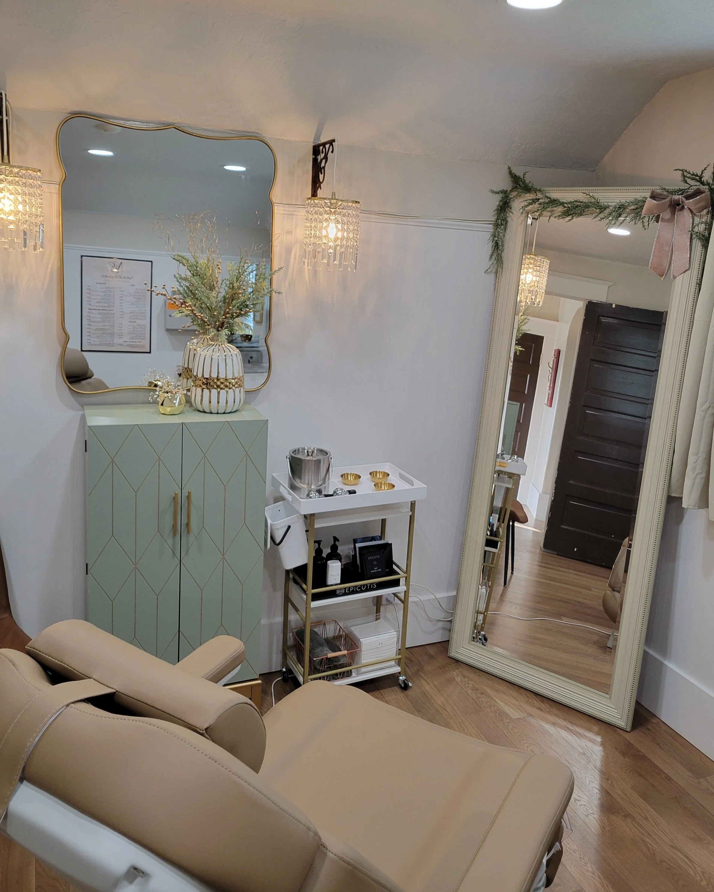 A cozy, well-decorated room with a large mirror, a cream-colored reclining chair, a small gold and white cabinet with a vase of greenery, a cart with skincare products, and an elegant mirror with festive greenery and a pink bow on the top corner.