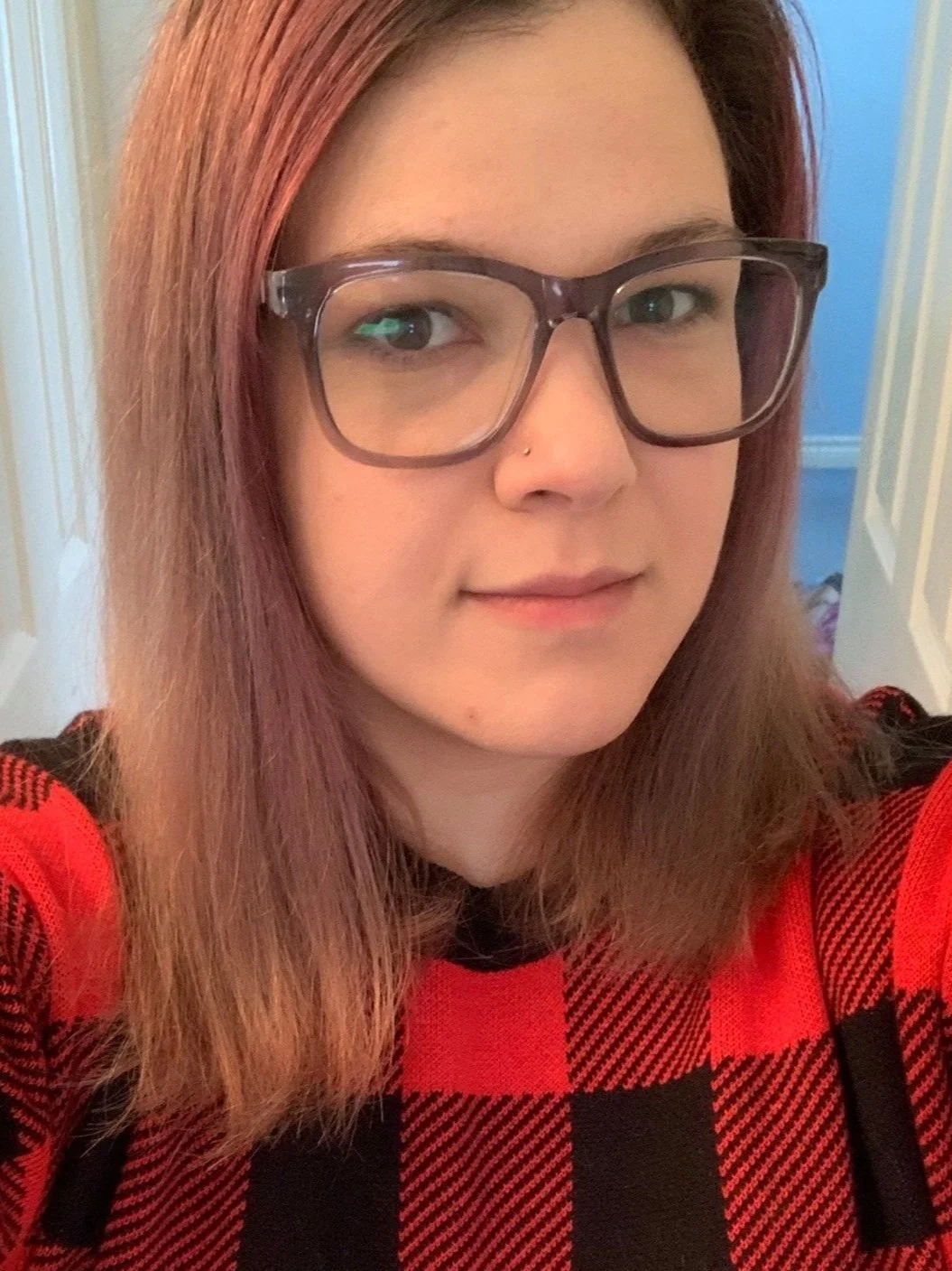 A woman with red hair, glasses, a small nose piercing, wearing a red and black checkered shirt, standing indoors with a doorway and light-colored walls in the background.