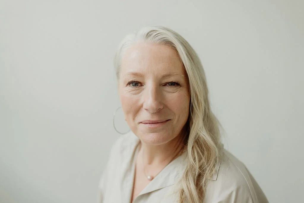 A portrait of a middle-aged woman with shoulder-length blond hair, light skin, and a subtle smile, standing against a plain light background.