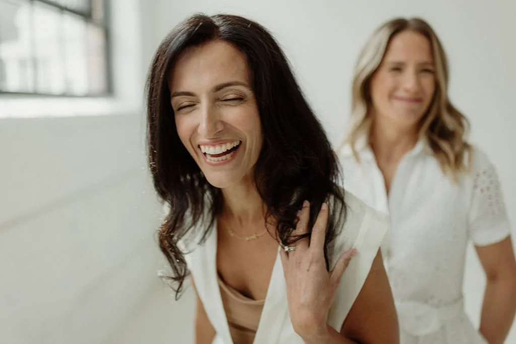 Two women smiling, one with dark hair in the foreground and one with blonde hair in the background, both dressed in white, in a bright room.