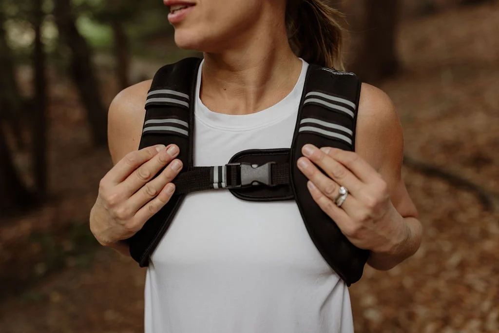 A woman wearing a white tank top and a black backpack with gray straps, standing in a wooded outdoor area.