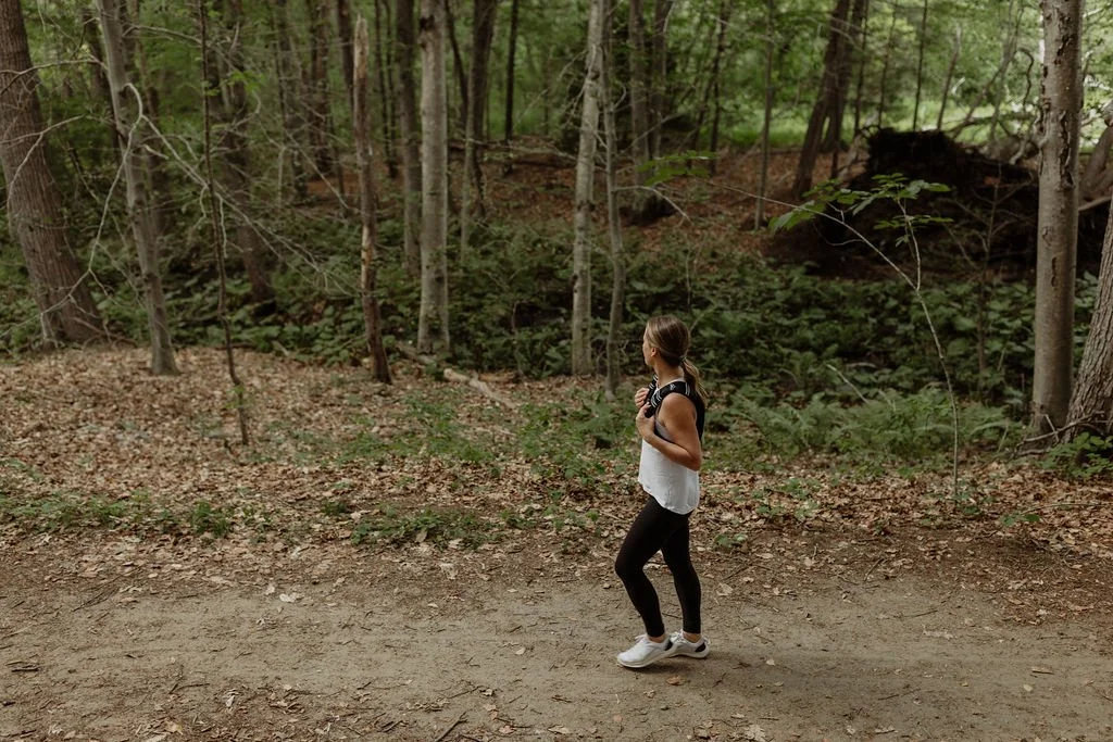 A woman walking alone on a dirt trail in a dense forest, surrounded by trees and green foliage.