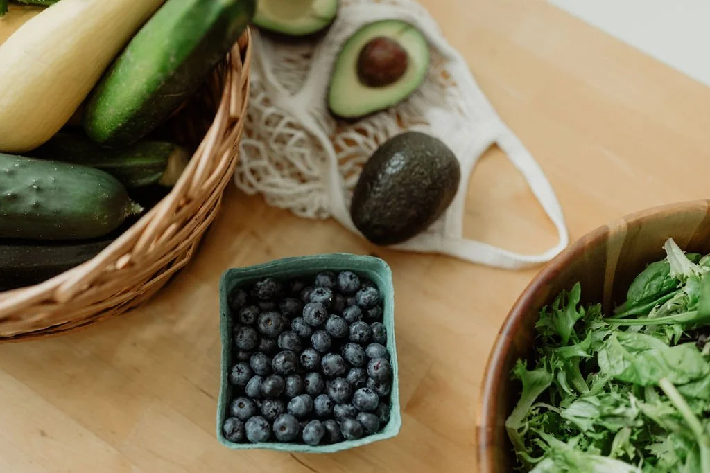 A basket of cucumbers and zucchinis, a small container of blueberries, and a wooden bowl of leafy greens on a light wooden surface, with an avocado and a cut avocado with the pit visible, and a white crocheted face mask with avocado illustrations in the background.