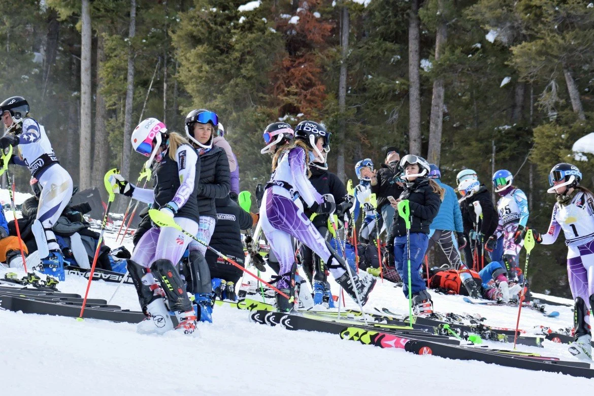 Alpine ski racers gathered with skis and poles beside a snowy forest on the Kirkwood Ski Education Foundation full-time program page