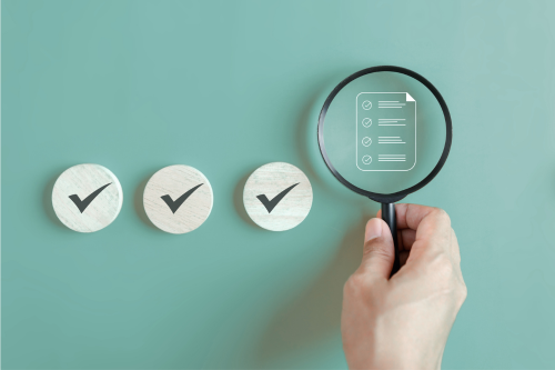 Hand holding a magnifying glass over three checkmarks on circular papers arranged in a row on a teal background.
