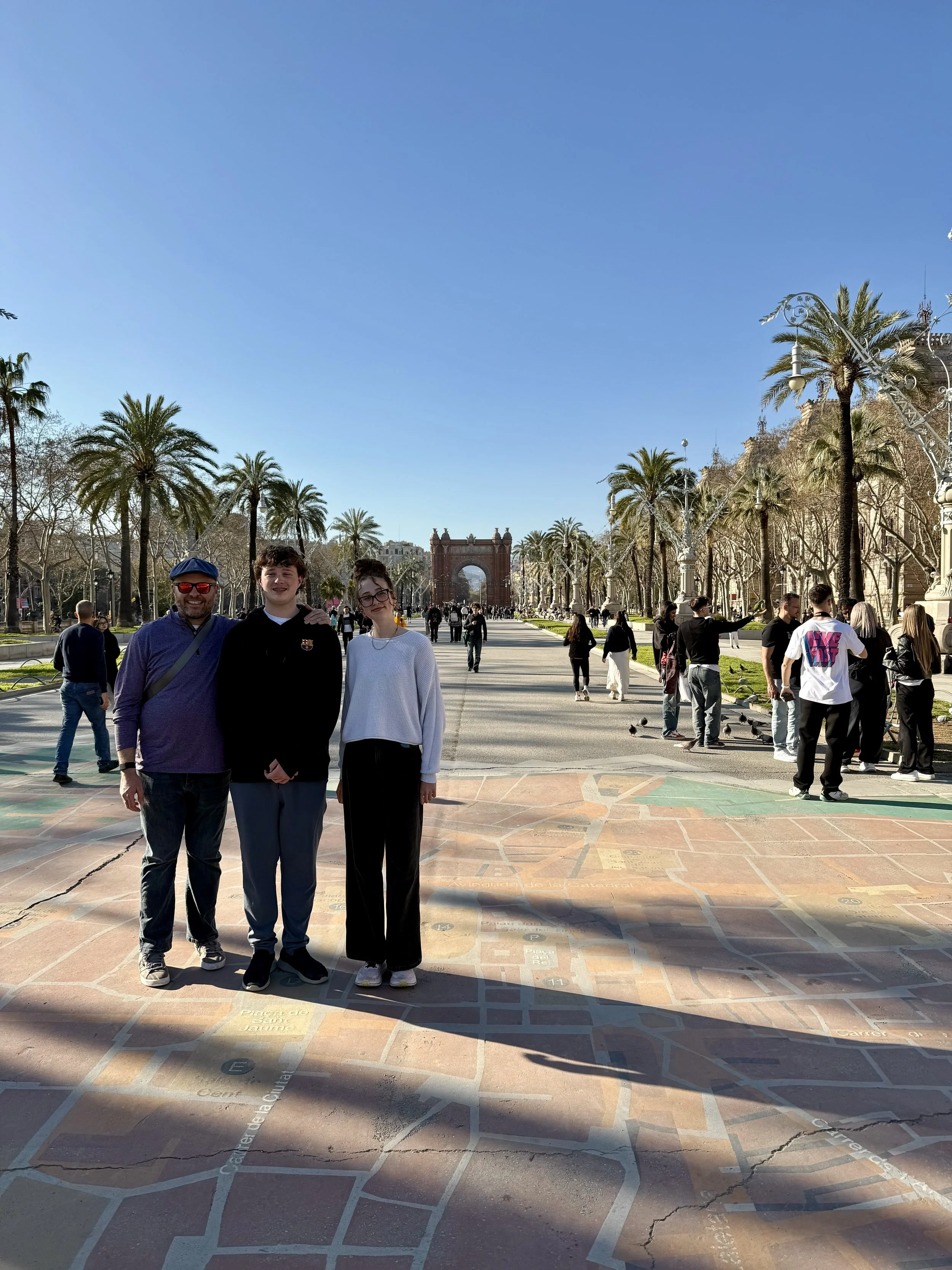 Three people posing in front of Arc de Triomf with palm-lined promenade