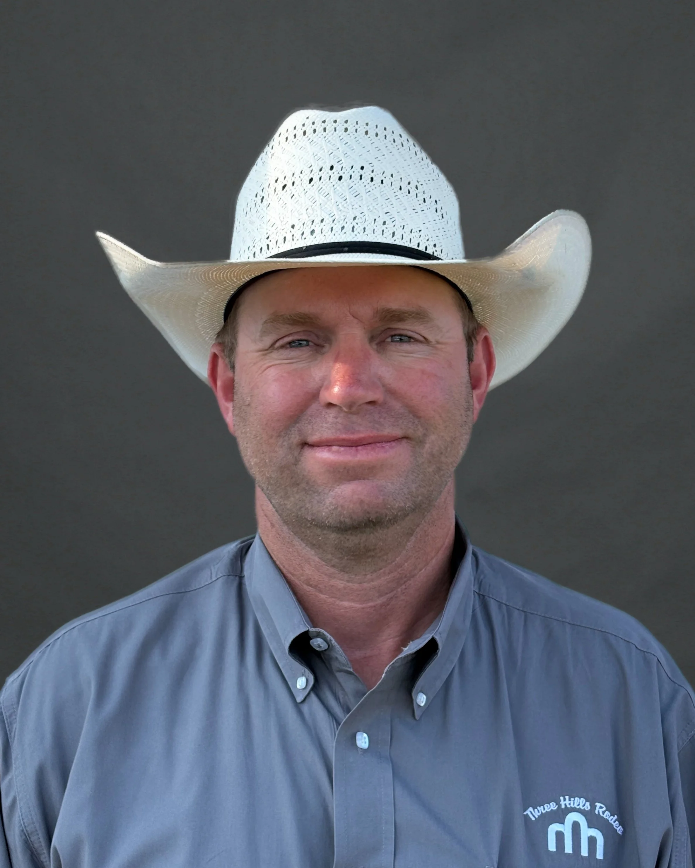 A smiling man wearing glasses, a black cowboy hat, a black jacket, and a dark shirt against a dark background.