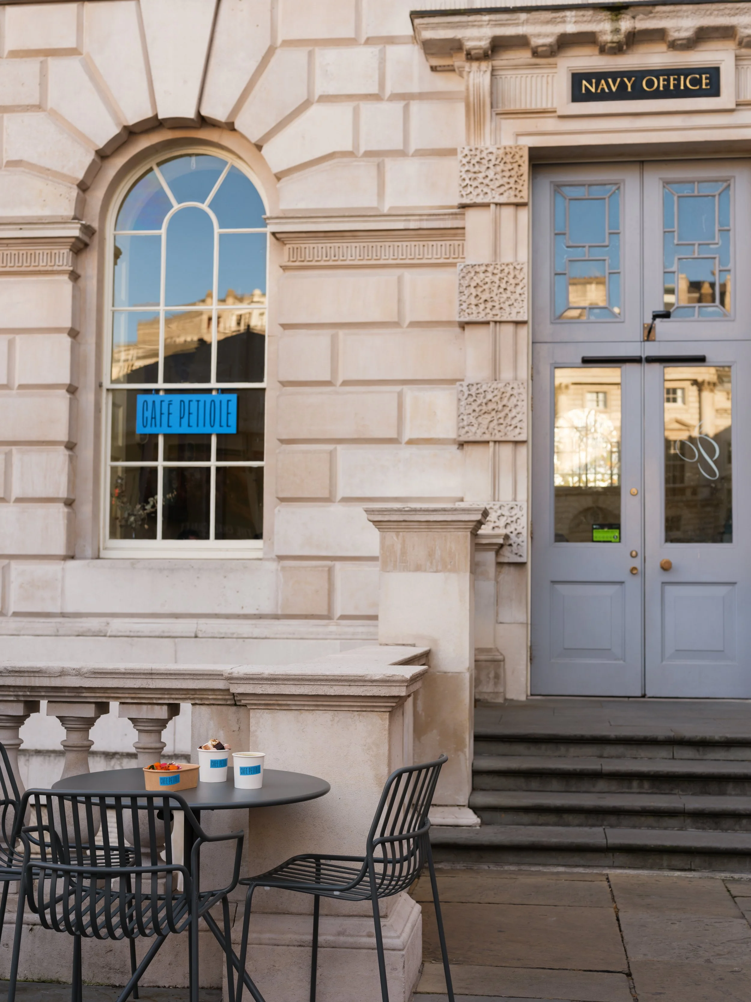 Outdoor scene with black metal table and chairs on sidewalk in front of a building with stone facade, arched window, and a door with a sign saying 'Navy Office'. The table has cups and an ice cream container from Cafe Petiole.