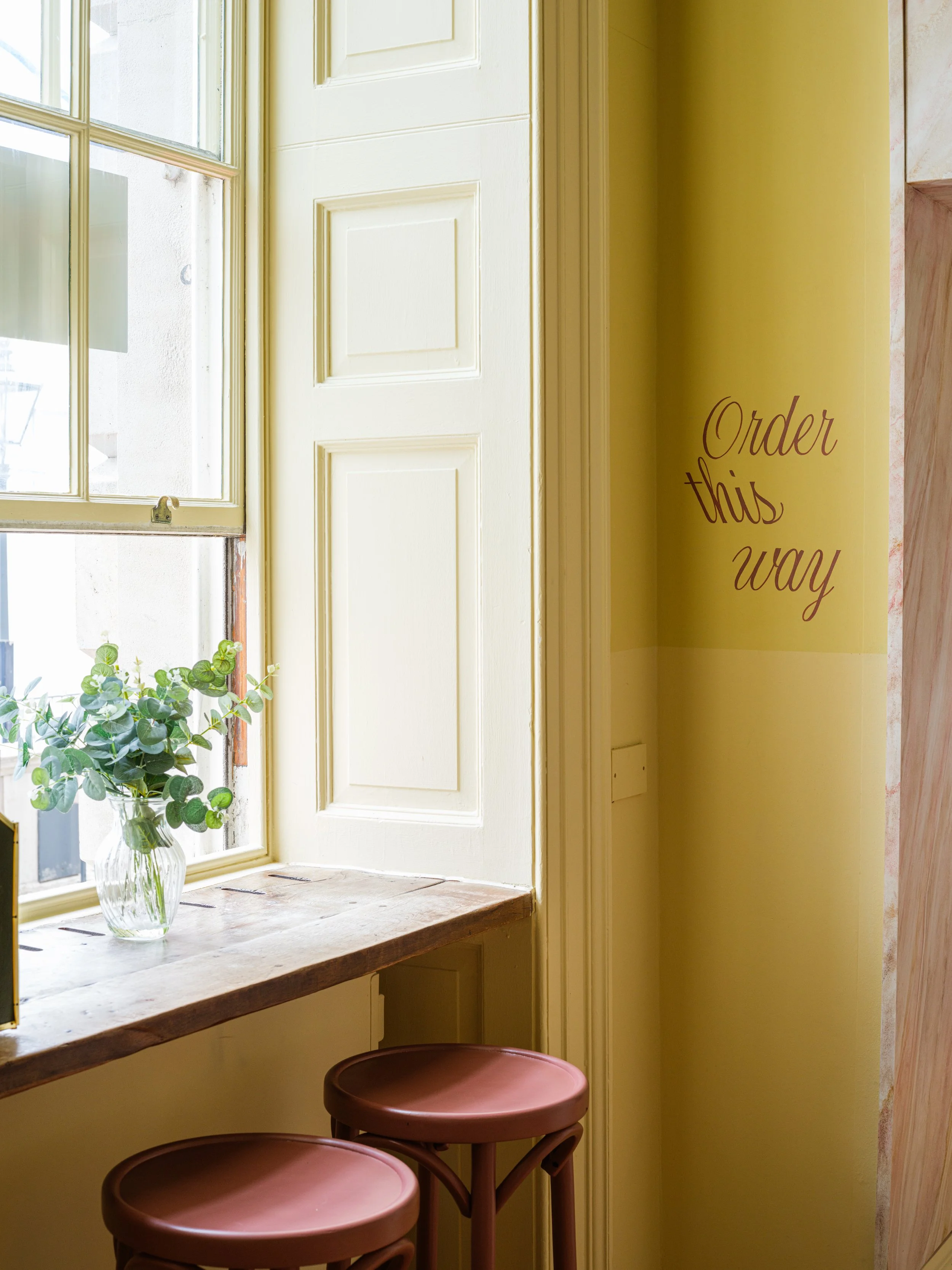 A cozy Cafe Petiole interior with a large window, a wooden window seat, a yellow wall with a handwritten sign that says "Order this way," and two pink stools beneath the counter.
