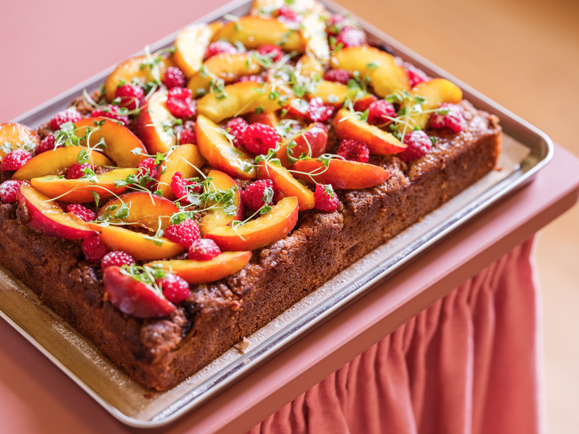 Cafe Petiole fruit-topped dessert with a brown crust, garnished with sliced peaches, raspberries, microgreens, and a dusting of powdered sugar, on a silver tray.