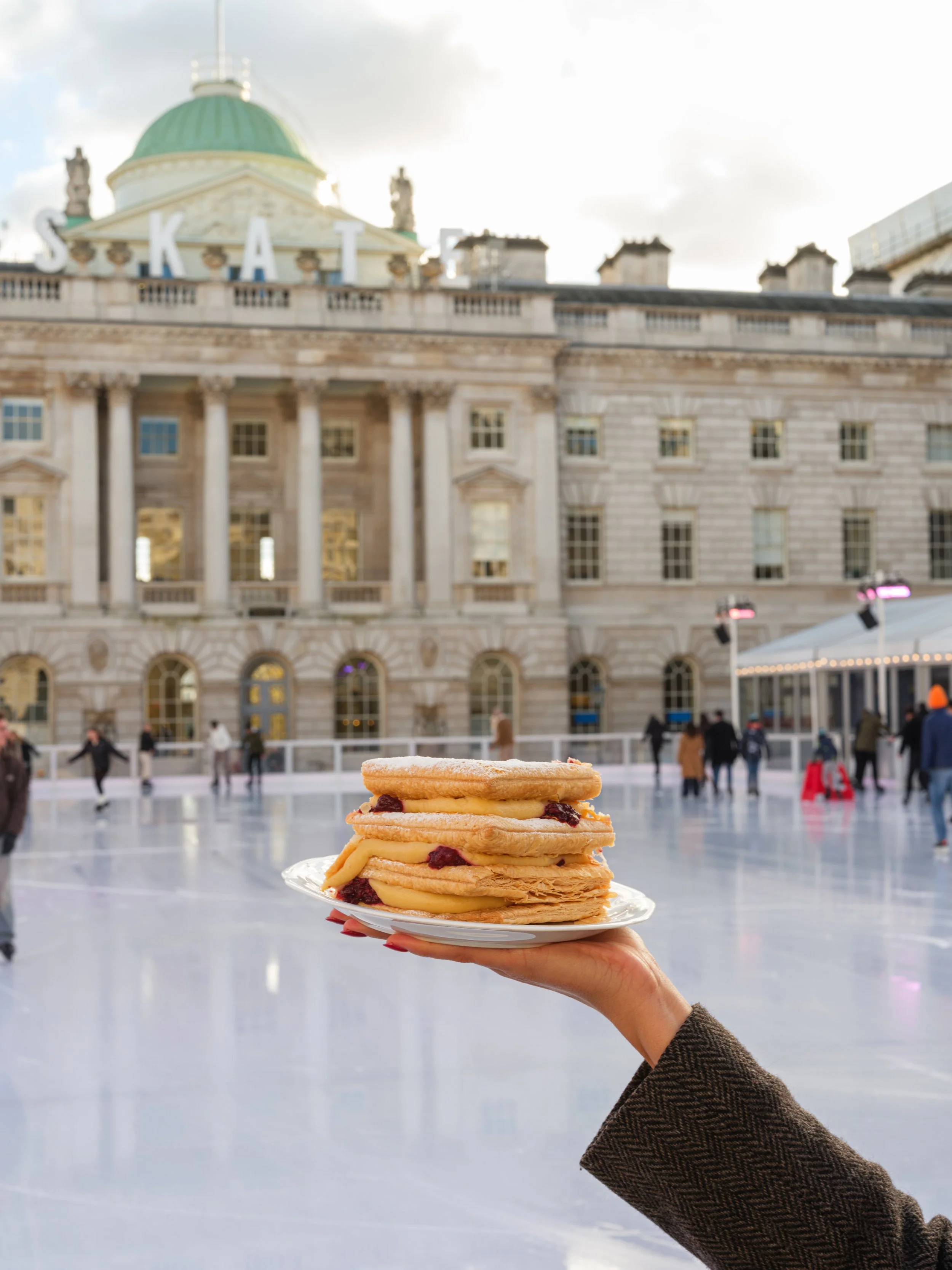 A hand holding a plate of Cafe Petiole cake in front of an ice skating rink with a historic building and a sign that reads 'SKATE' in the background at Somerset House.
