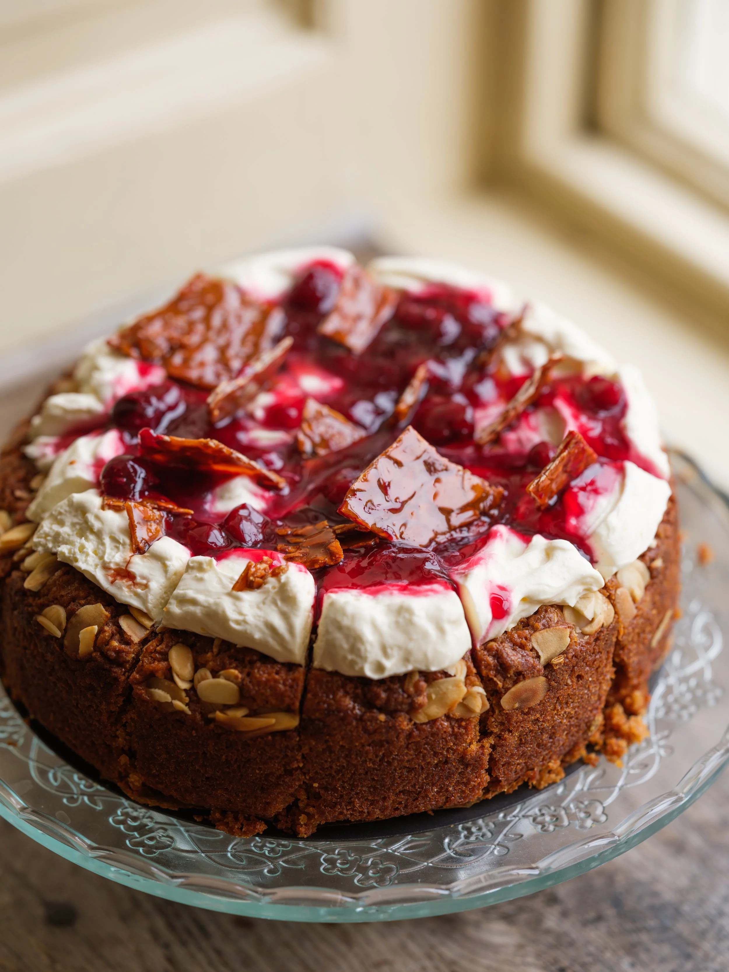 A close-up of a decorated cake with whipped cream, cherry topping, and chopped nuts on the sides, placed on a glass cake stand.