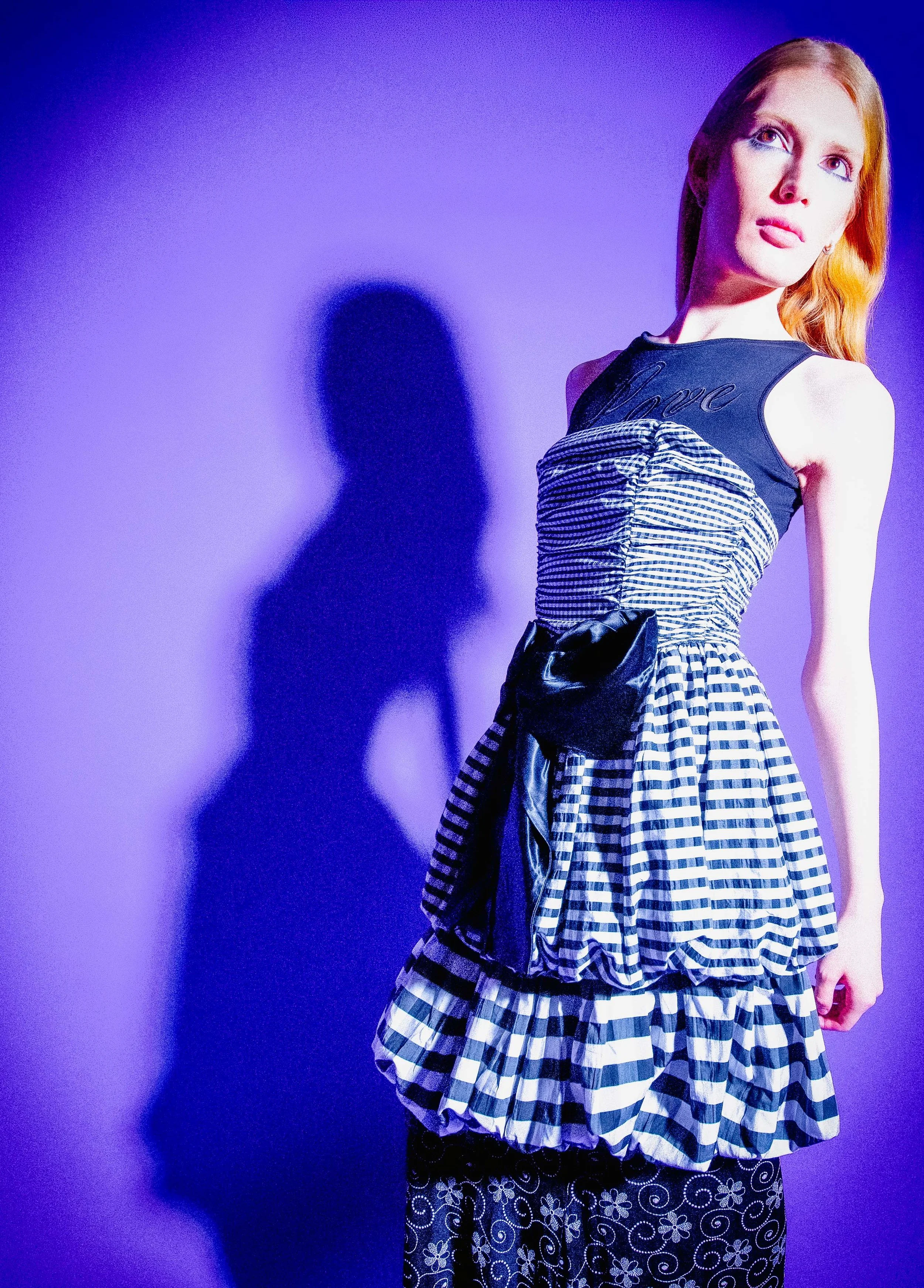 Strawberry blonde model in a gingham ruffle dress with a black bow, against a violet backdrop. Vintáááge June by Hartenjager.