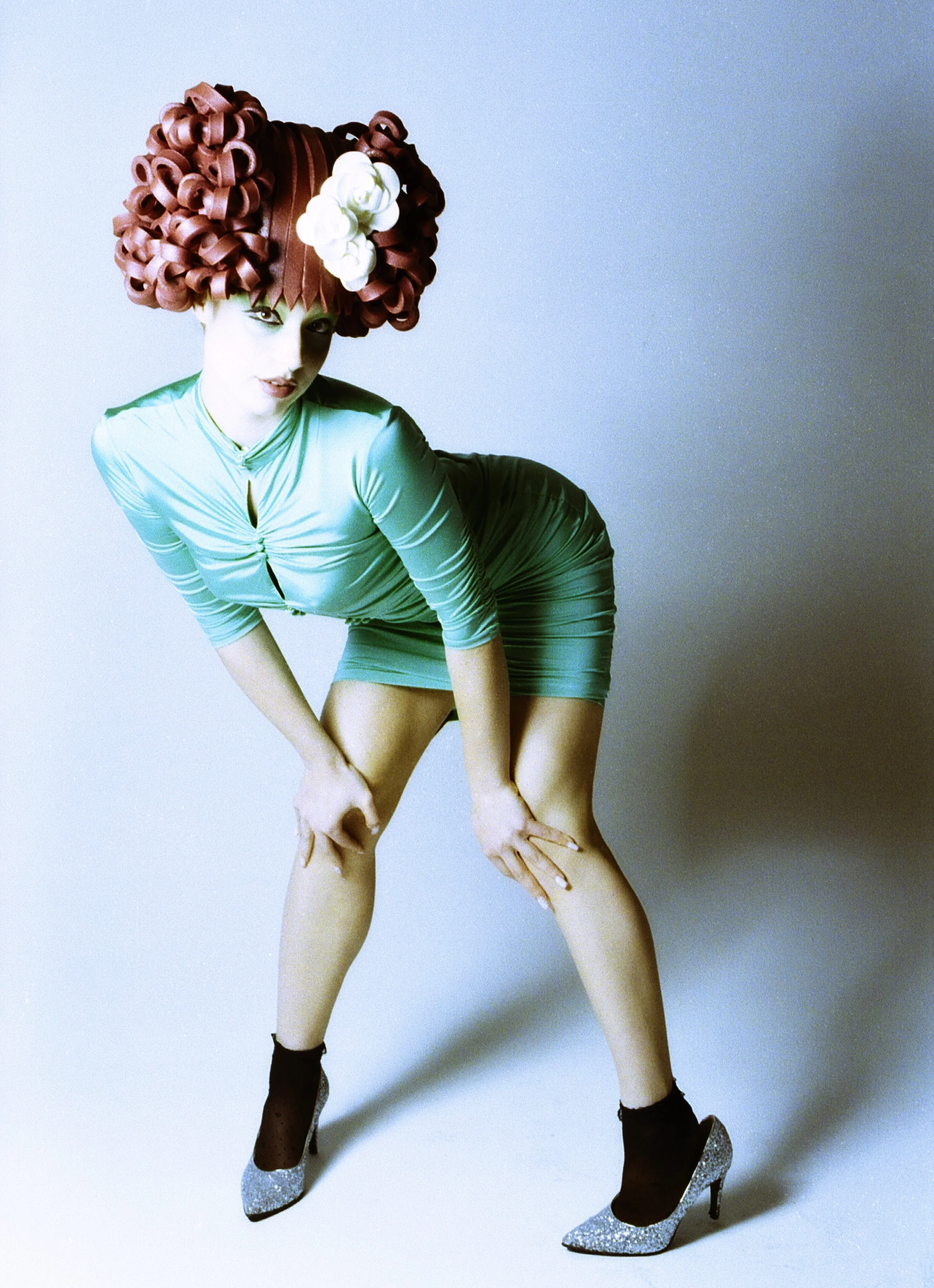 Model in a teal two-piece and sculpted red curl wig bending forward in heels. Loopsyland, editorial for Kaltblut Magazine by Hartenjager.