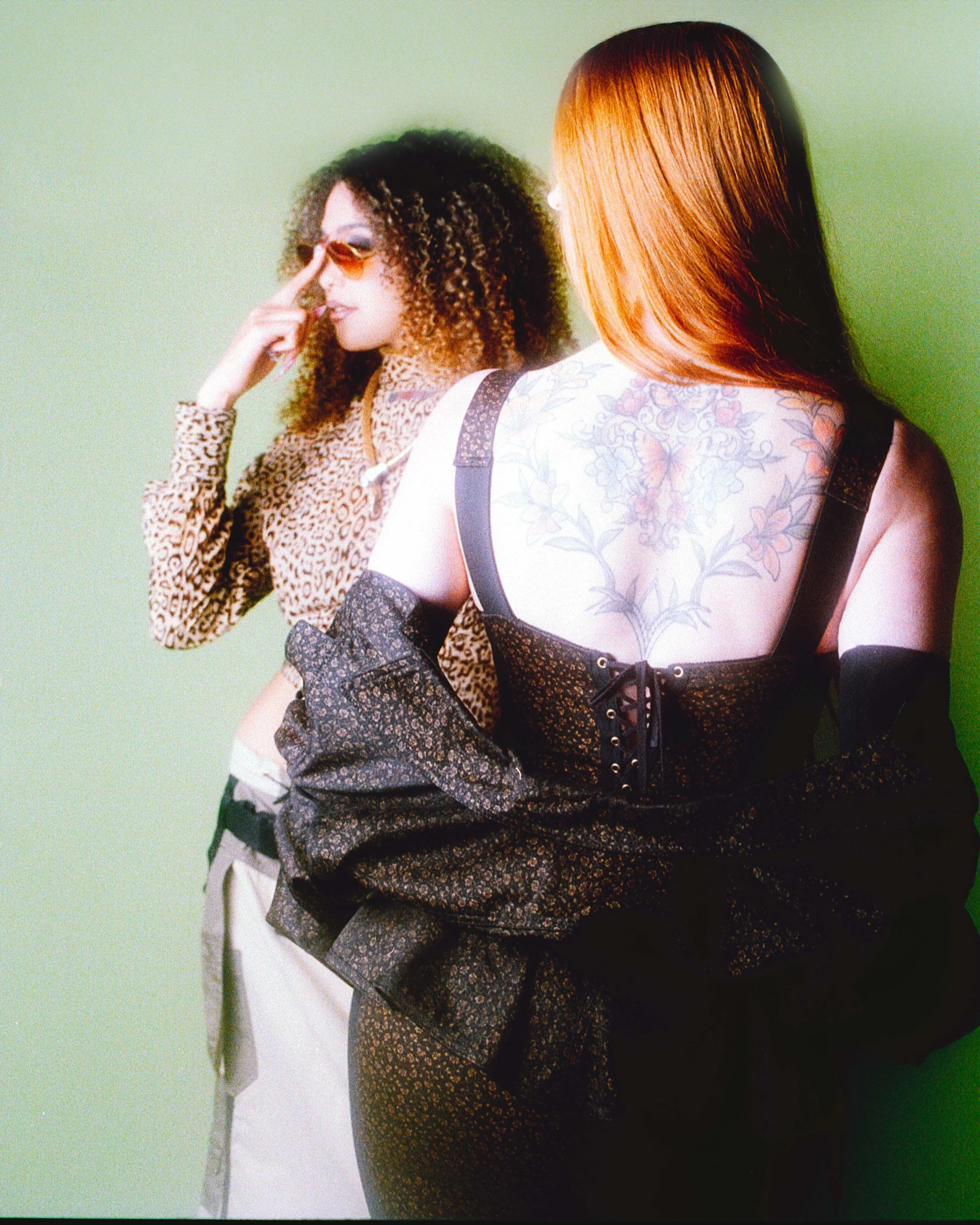 Two models back to back against green, one in sunglasses and leopard print, one showing a back tattoo. Vintáááge May by Hartenjager.