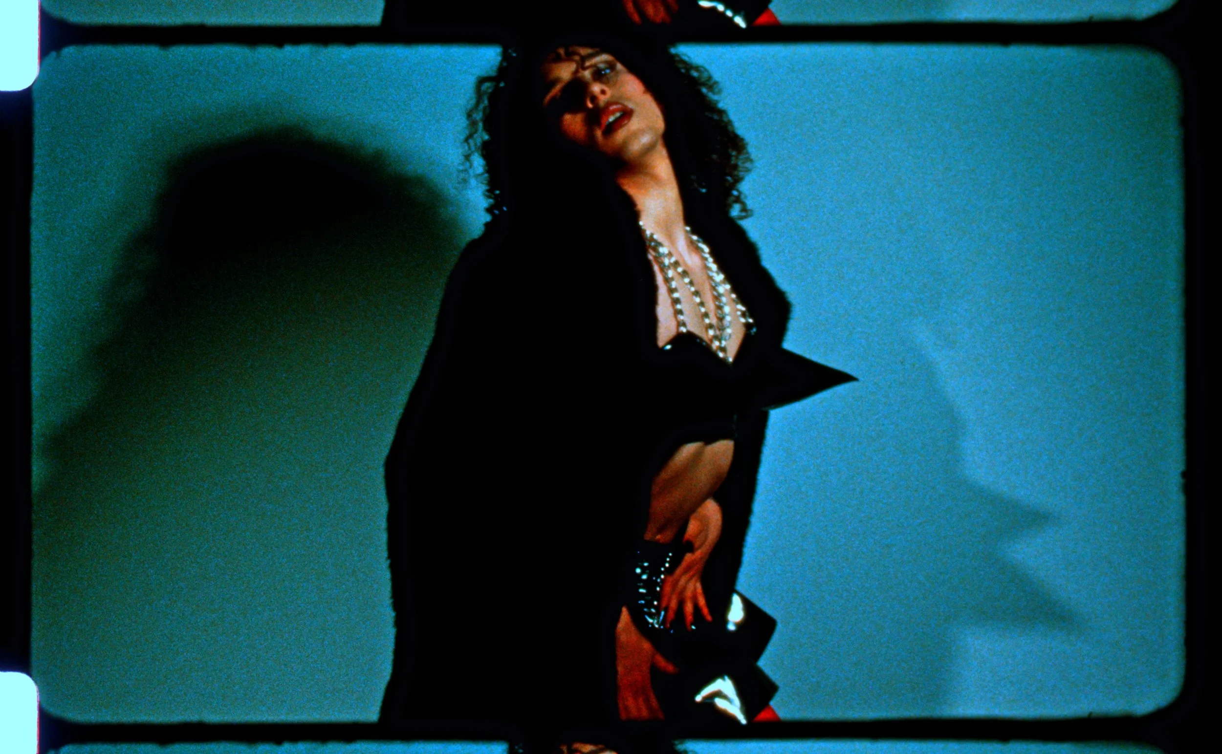 16mm frame of a figure in a long black coat standing in blue light. Still from Intrusive Thots, 16mm film by Hartenjager.