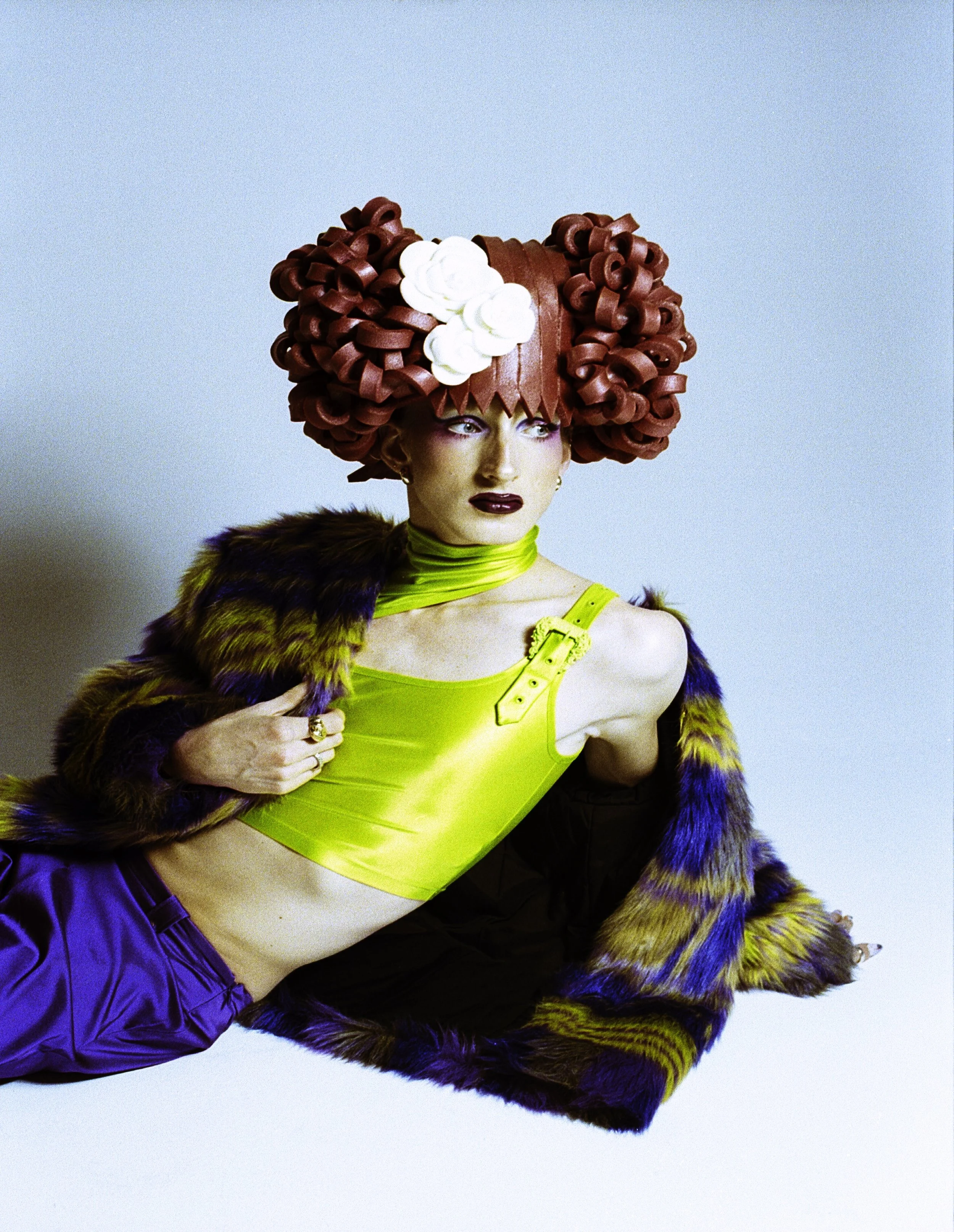 Model in a sculpted red bun wig and neon yellow top lying across a patterned coat. Loopsyland, editorial for Kaltblut Magazine by Hartenjager.