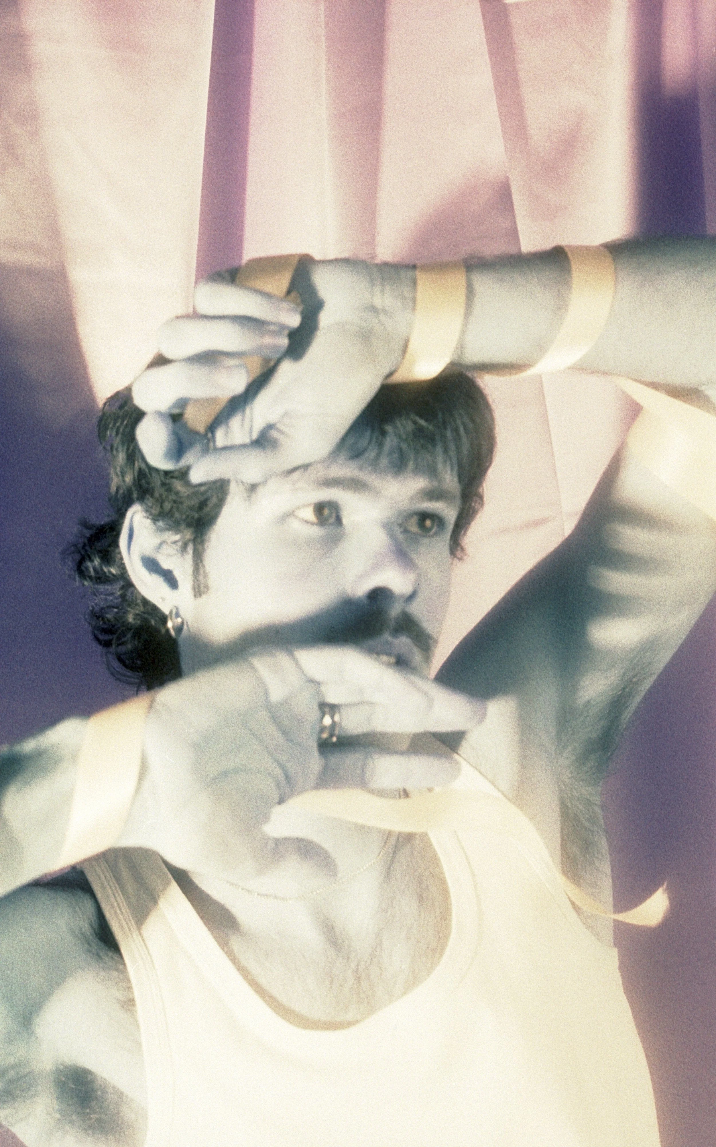 Double exposed film portrait of a man with dark curly hair and a moustache, hands over his face. By Hartenjager.