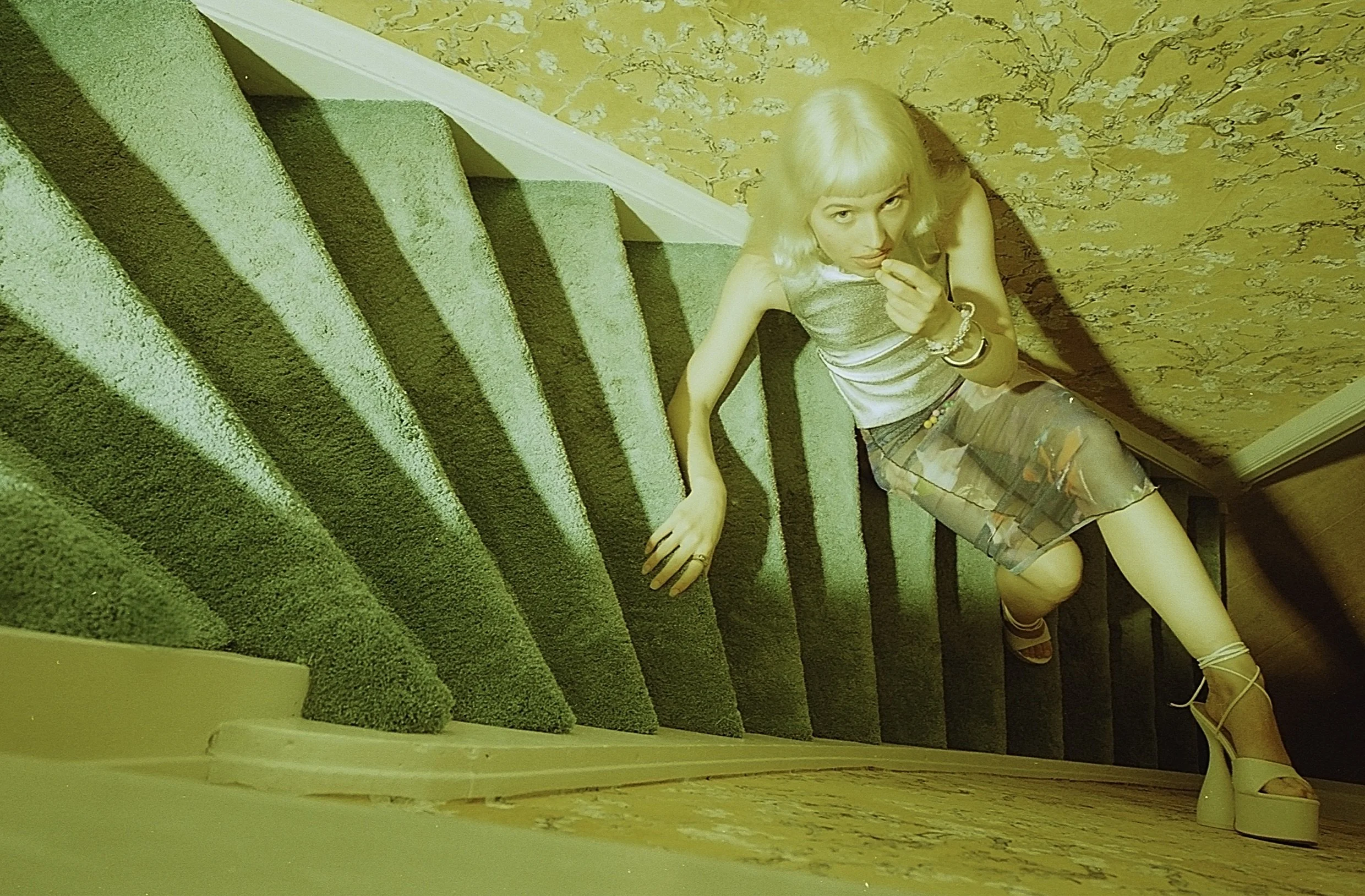 Blonde model in silver climbing down a steep wooden staircase in warm yellow light. Bubblehead by Hartenjager.