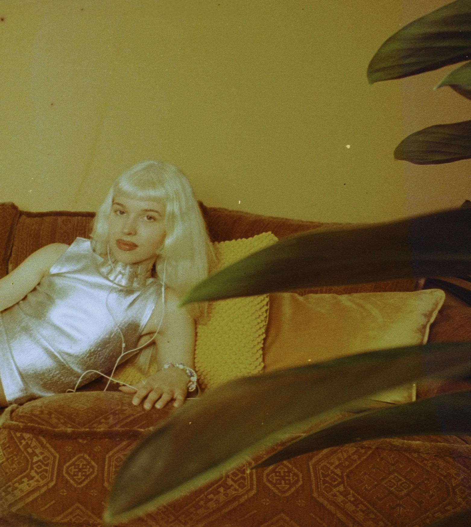 Blonde model in a silver satin dress lounging on an orange velvet sofa behind large green leaves. Bubblehead by Hartenjager.