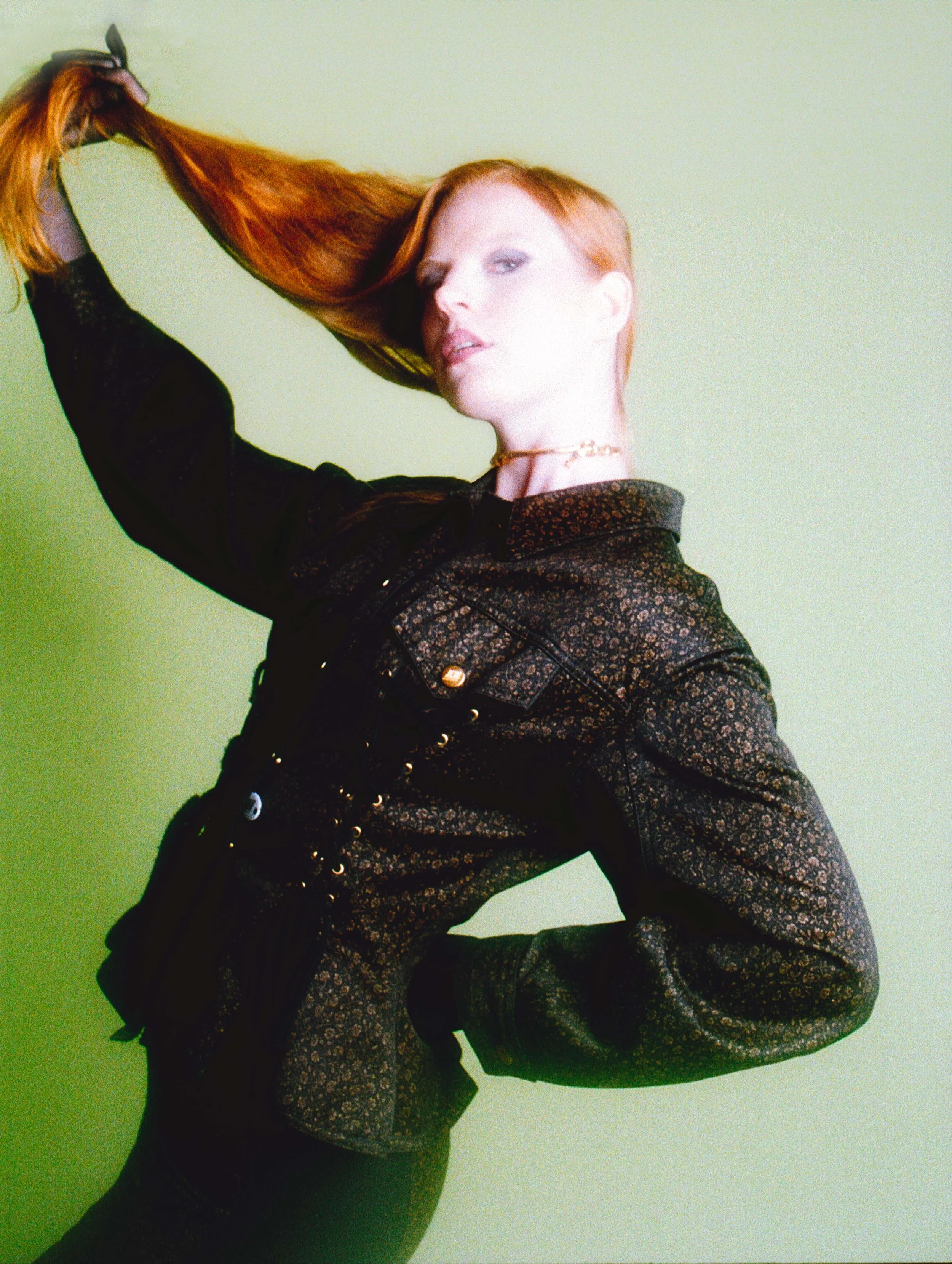 Red-haired model in a black jacket pulling her long hair up into the air against green. Vintáááge May by Hartenjager.