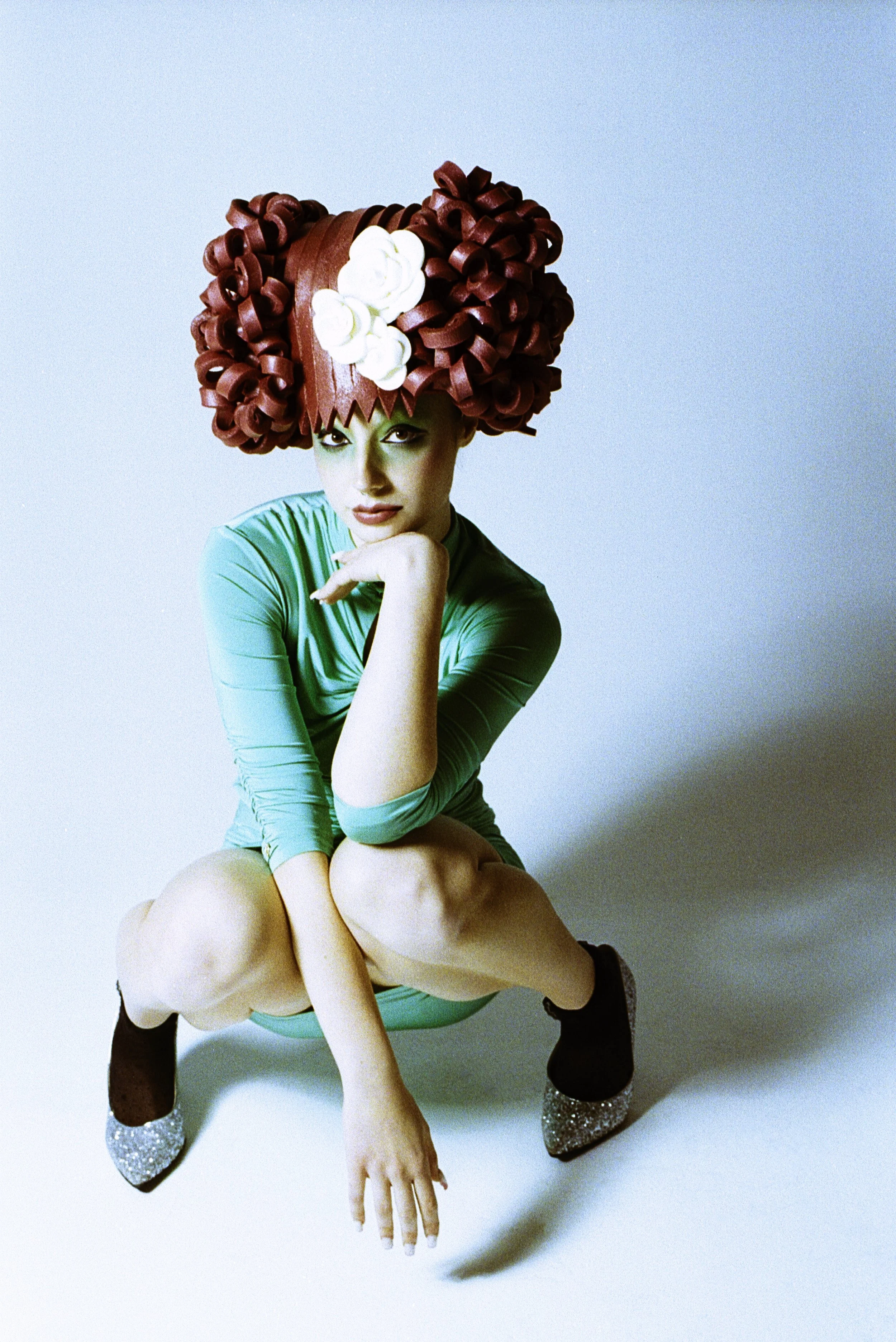 Model in a green bodysuit and a sculpted red curl wig crouching with chin in hand. Loopsyland, editorial for Kaltblut Magazine by Hartenjager.