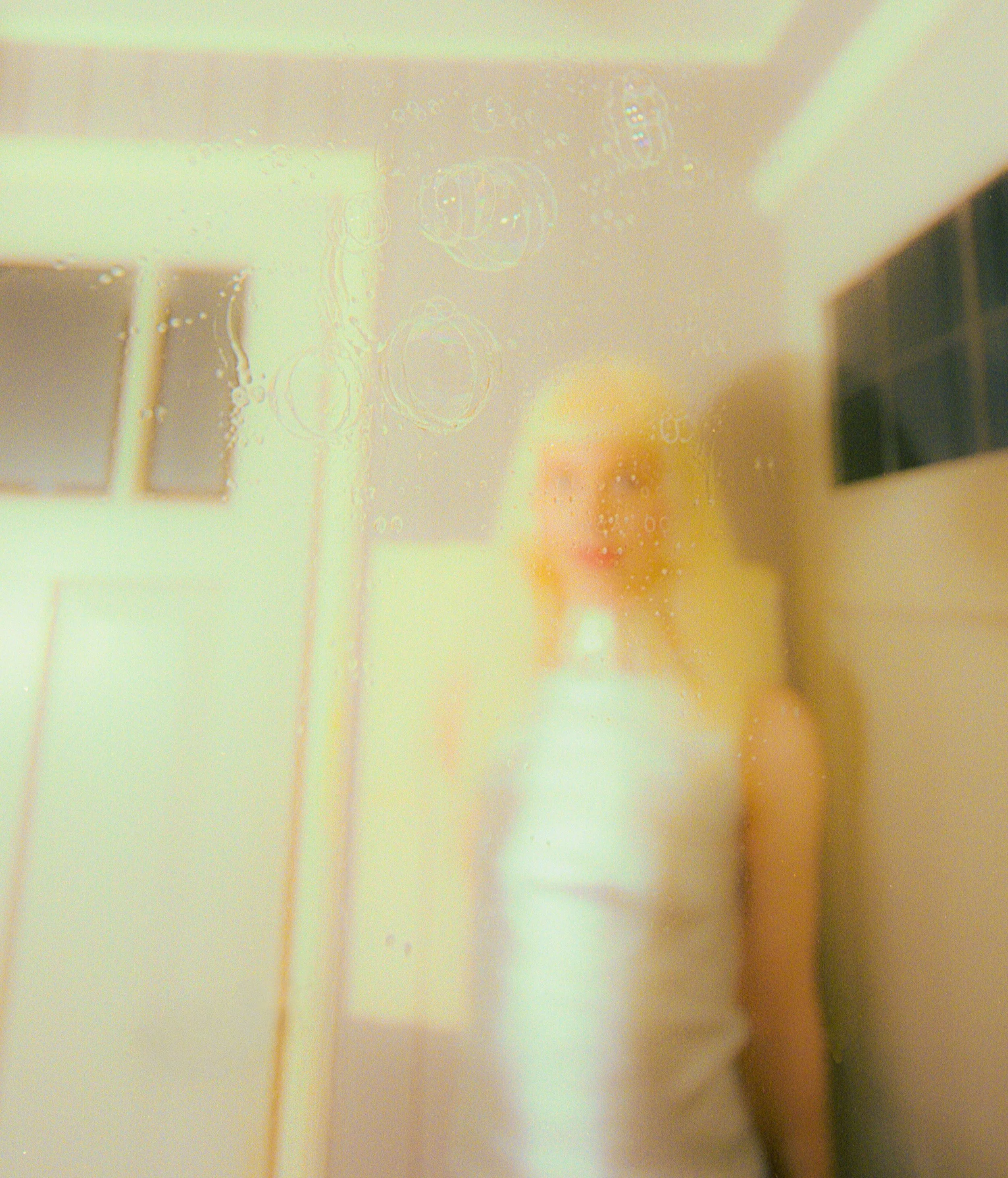Blurred, light-leaked film image of a blonde figure in a white top standing in a pale room. Bubblehead by Hartenjager.